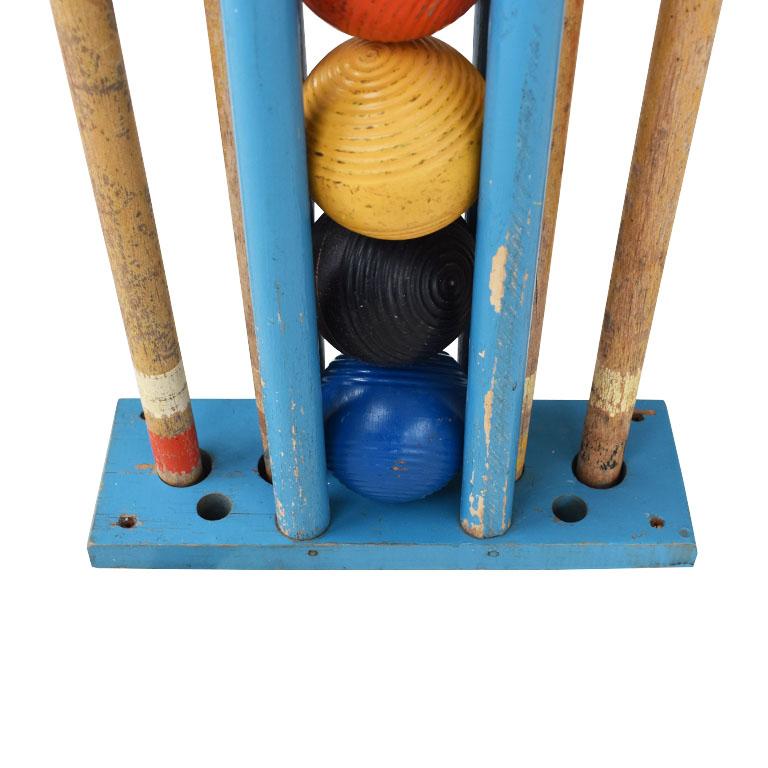 A lovely painted antique croquet set. This set includes four mallets, four balls, and metal stakes all in a wooden stand. This set is quite old and accommodates four players. The balls are painted orange, yellow, black, and blue. Each ball