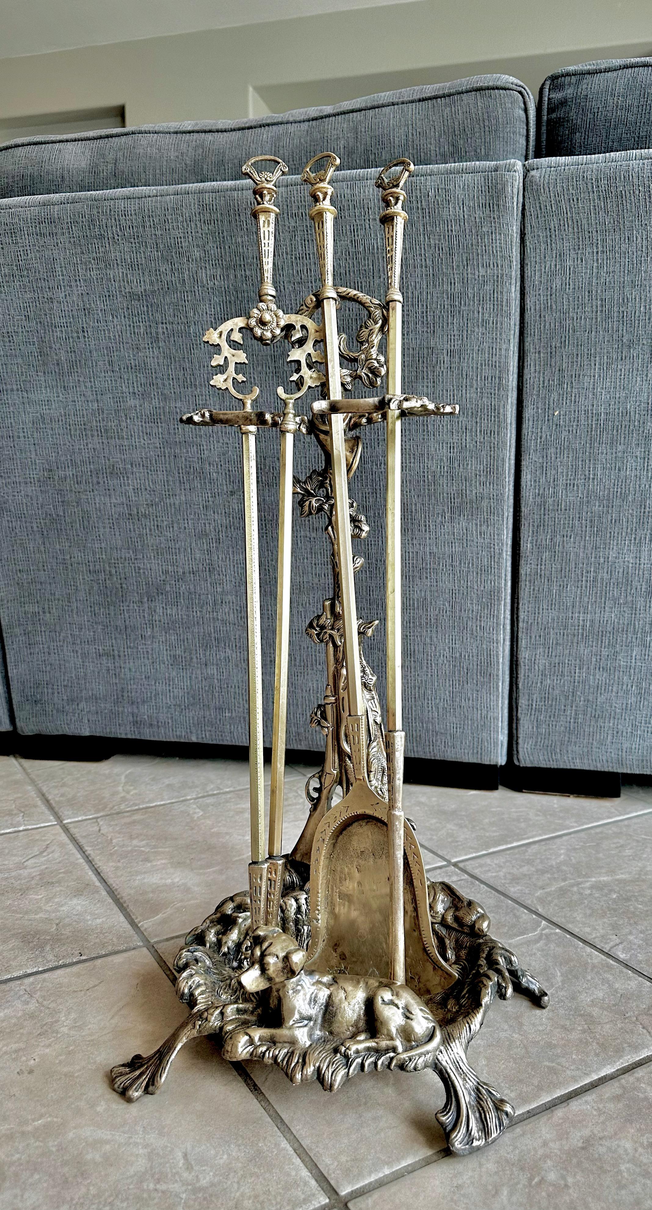 An antique hunting scene set of cast brass fireplace tools. The set has four total pieces; the base, a poker, shovel and grabbing piece. The hunting motif scene includes a dog sitting at the base, a gun, horn at the top, foliage some other animals.