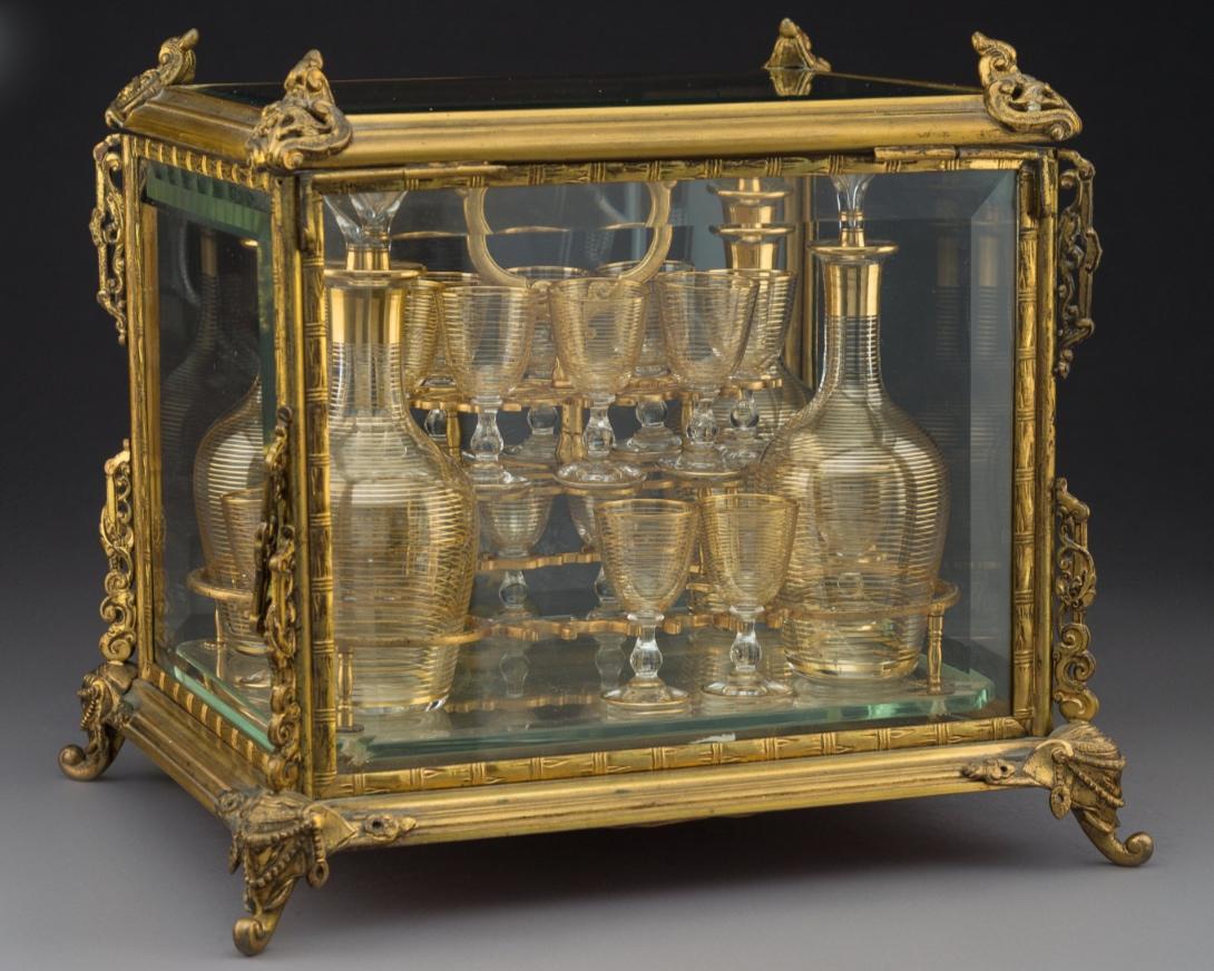 Our antique bronze and glass tantalus with four decanters with stoppers and fifteen glasses with raised gilt decorations measures 12 x 13.75 x 11.5 inches (30.5 x 34.9 x 29.2 cm). One glass is missing. The underside is reinforced with cardboard.
