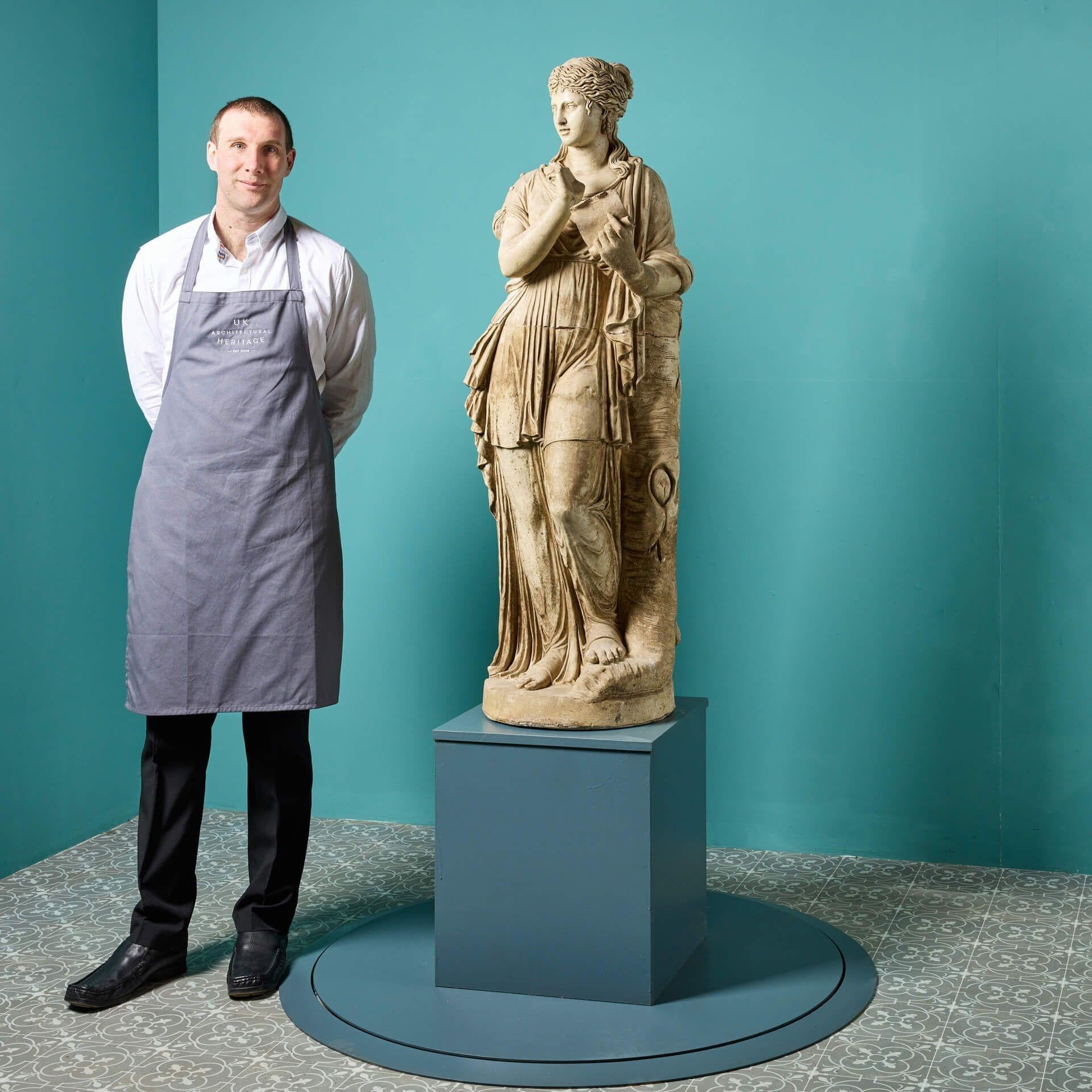 Eine antike französische Terrakotta-Statue in Dreiviertel-Lebensgröße von Clio, einer der 9 Musen der griechischen Mythologie, aus dem frühen 20. Jahrhundert. Clio, die Göttin der Geschichte, lehnt an einem Baumstumpf, ist in einen weiten Stoff