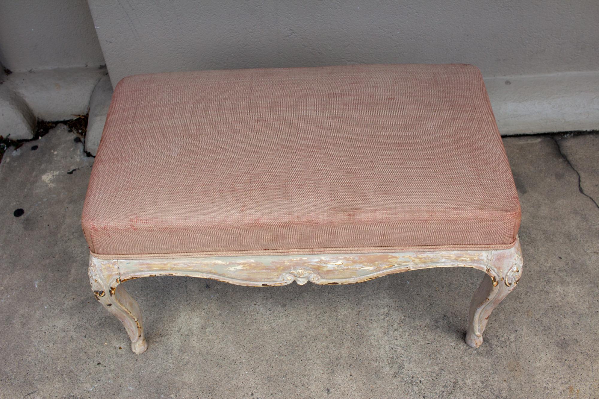 This is an antique French carved wood bench with dusty pink upholstery and a distressed painted finish. The decoration on the wood base includes Fleur de Lis shapes, scrolls and tapered, curved legs. The painted finish has tones of cream, grey and