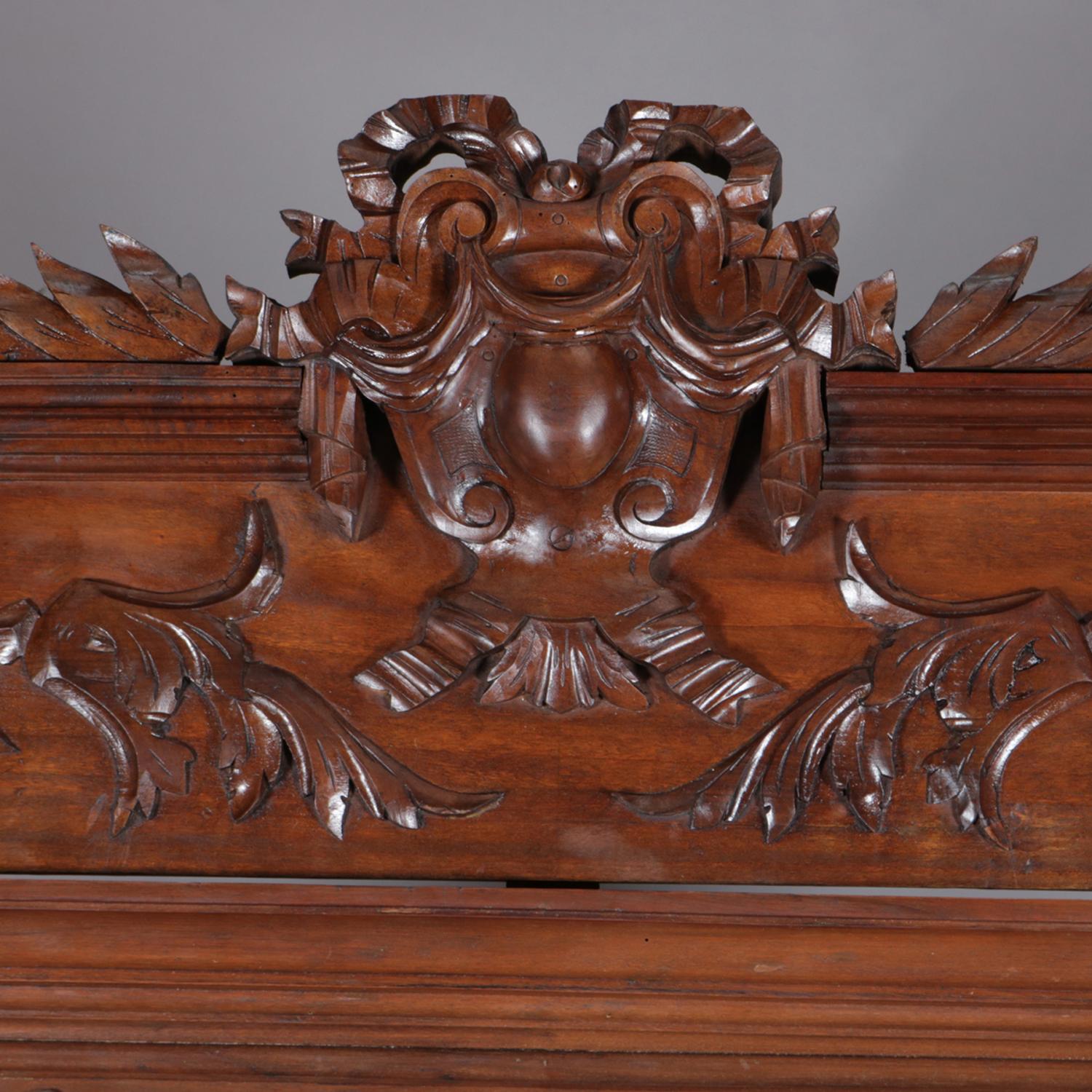 An antique French bookcase features walnut construction with broken arch crest having central carved shield, garland and foliate decoration surmounting case with two doors with arched and beveled glass opening to shelved interior and flanked by