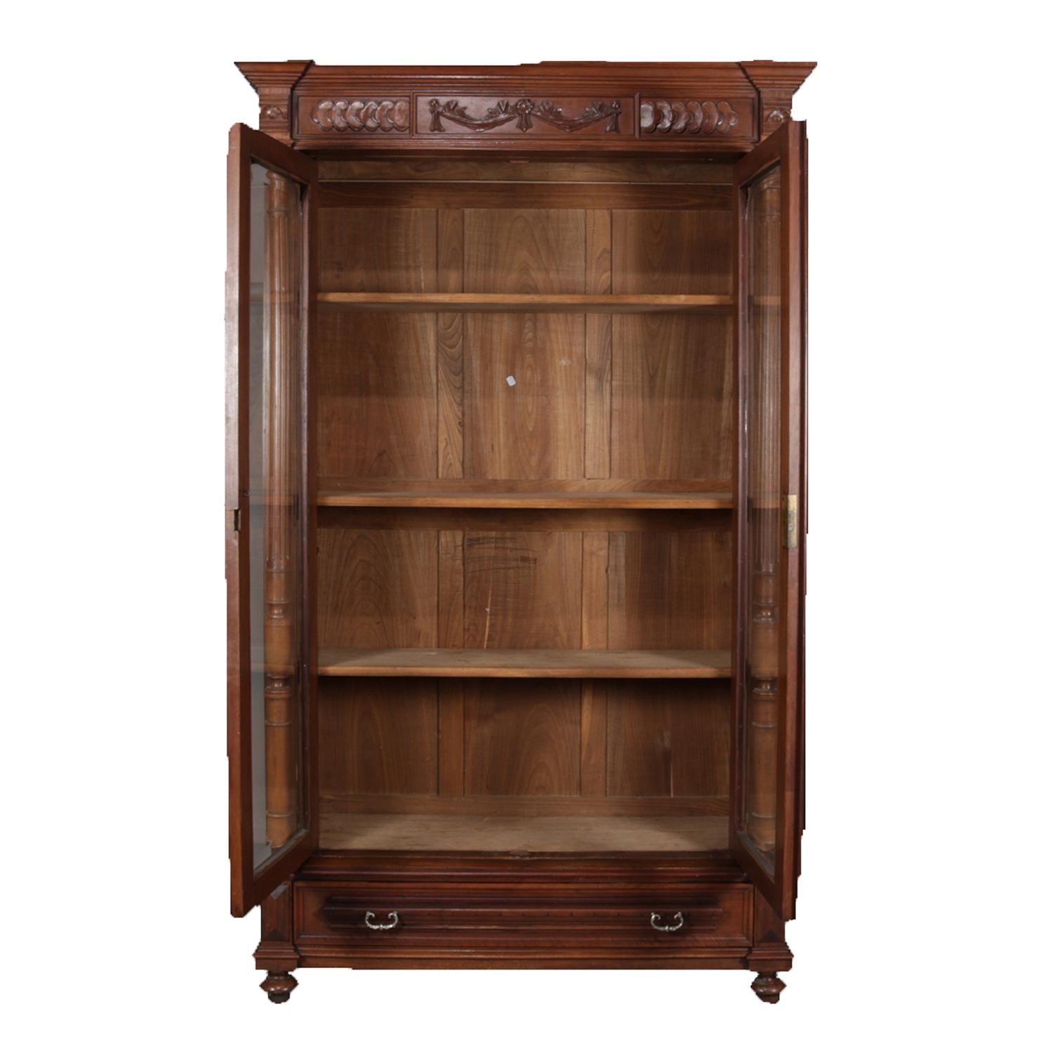 Antique French bookcase features walnut construction with crest having Lincoln drape carving over two glass doors opening to adjustable shelf interior and flanked by turned columns and over single long drawer and raised on turnip feet, circa