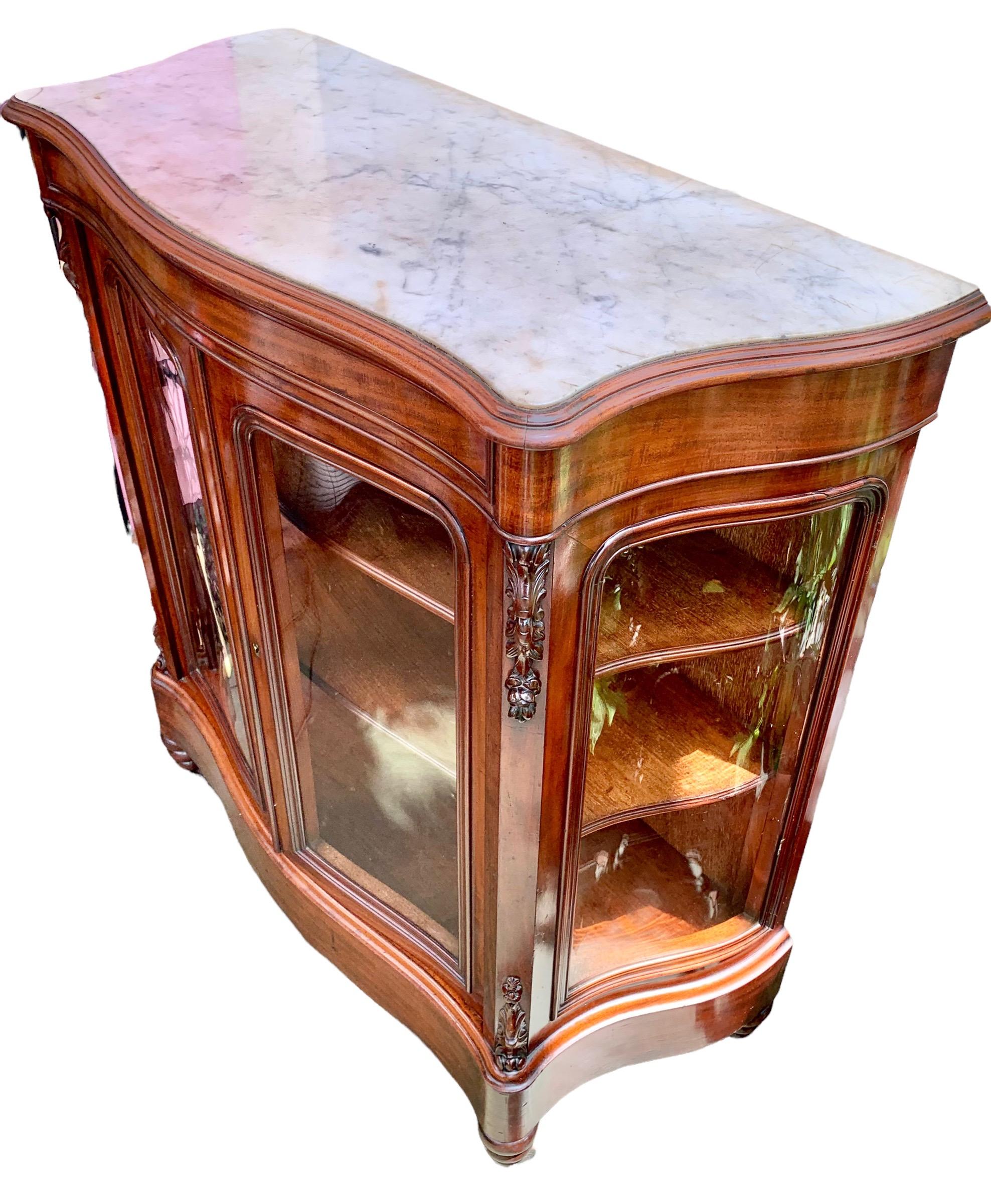 A French carved walnut marble top bowfront parlor cabinet, circa 1870, having a lovely inset white marble with double curved glass doors, flanked by curved glass sides. The plinth base above turned tapered legs with casters. The interior with two