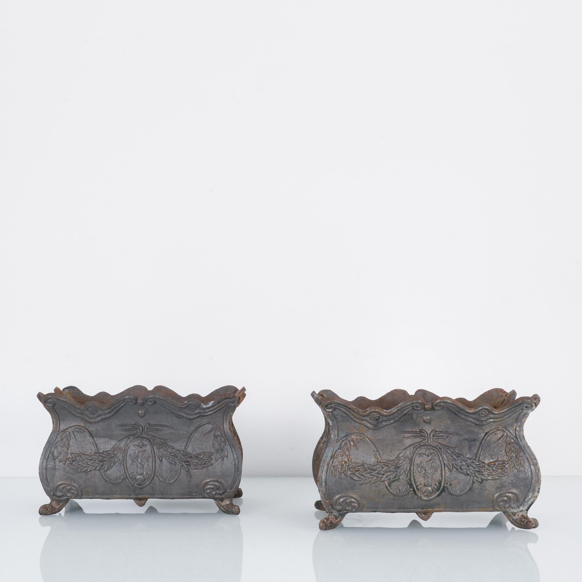 This pair of cast iron planters was made in France, circa 1920. The Art Deco influence is visible in its symmetry, stylized molding, and decorative references to nature. Raised on four scroll legs with a scalloped top, the slate gray planters