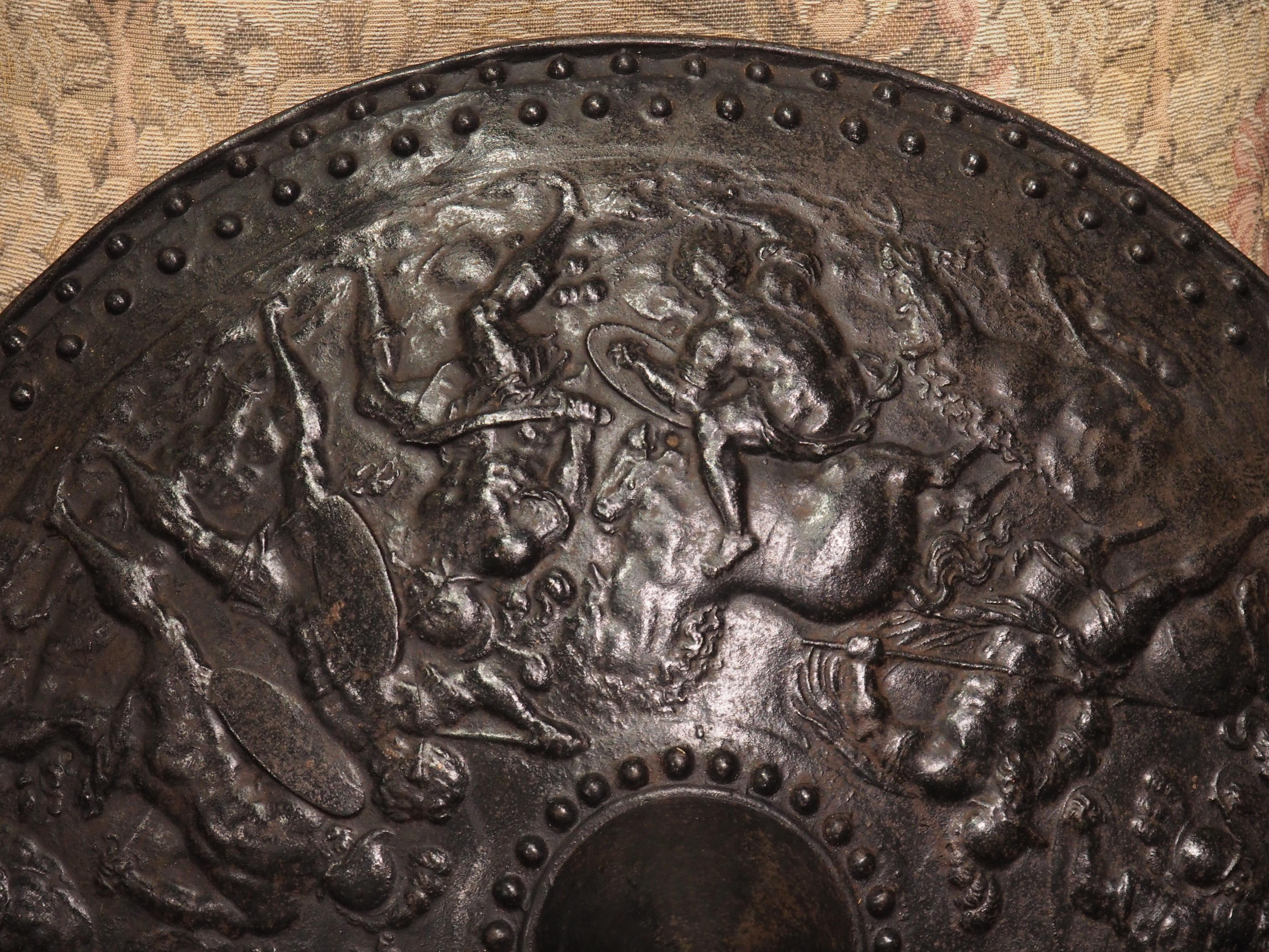 A fascinating iron shield that was cast in France, circa 1890, this round armorial piece features a central conical umbo (sometimes called a “shield boss”) surrounded by a 360-degree scene depicting a military engagement. The brutal skirmish depicts