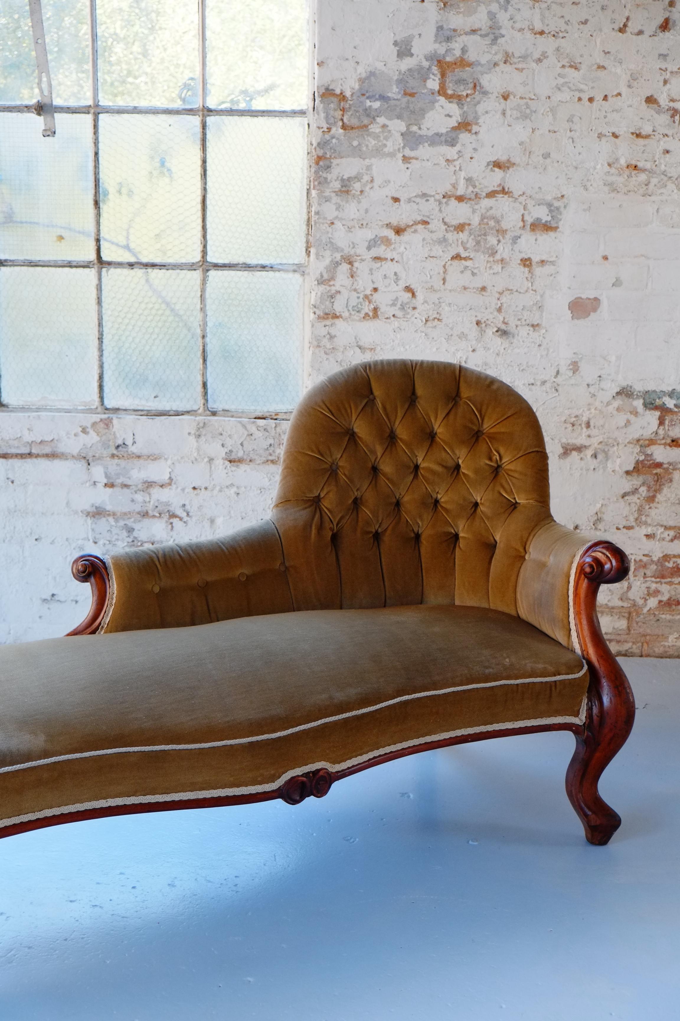 This is a handsome example of an antique French hand-carved chaise longue in Walnut. The chaise longue is upholstered in thick velvet, which is an earthy green, khaki color. The trimmings and piping are in creamy white to give the seat definition.