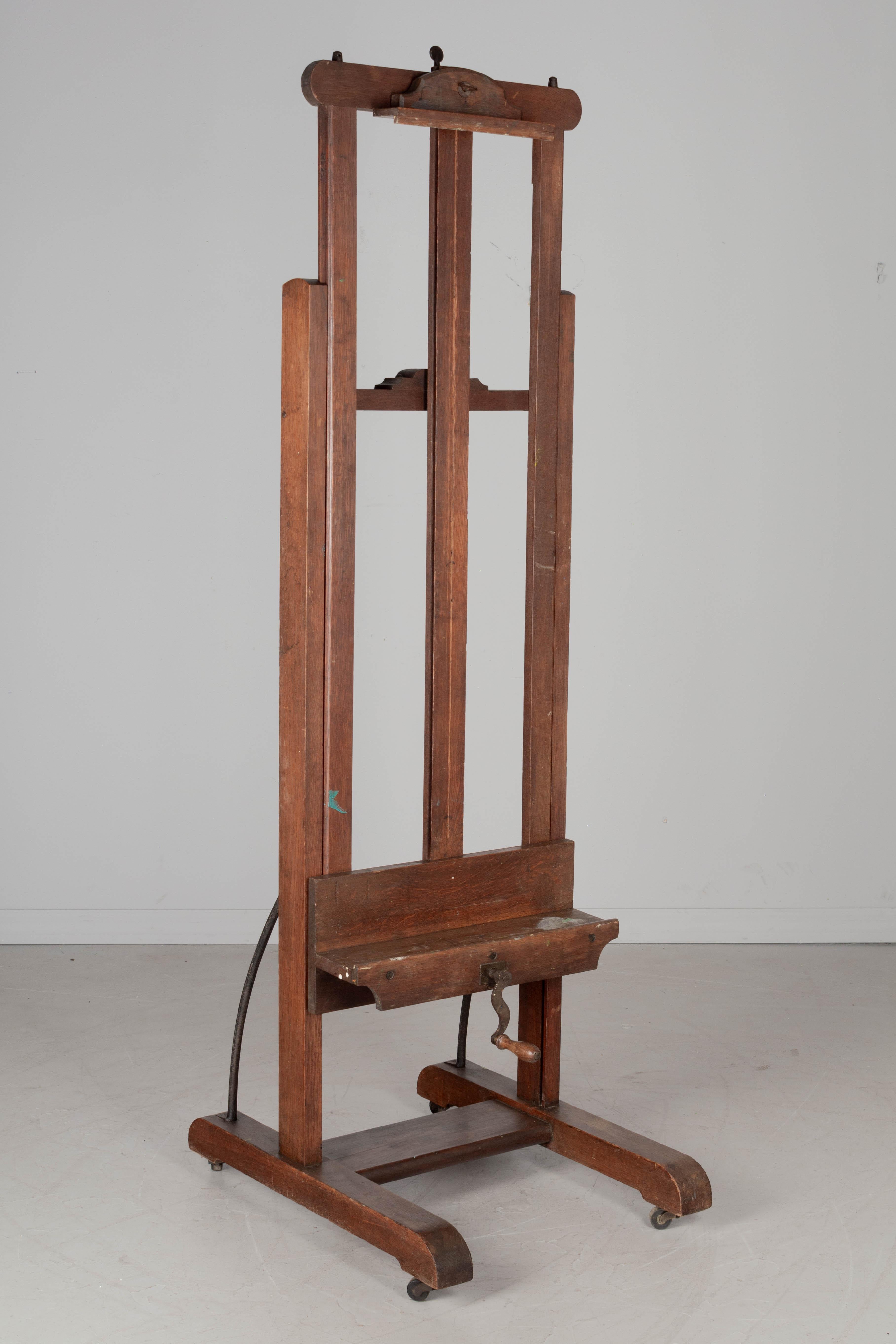 A large early 20th Century French painter's easel made of solid oak with threaded iron rod, cast iron gear and metal crank with turned wood handle. Ledge with remnants of spattered paint. Adjustable bracket at the top securely holds the painting or