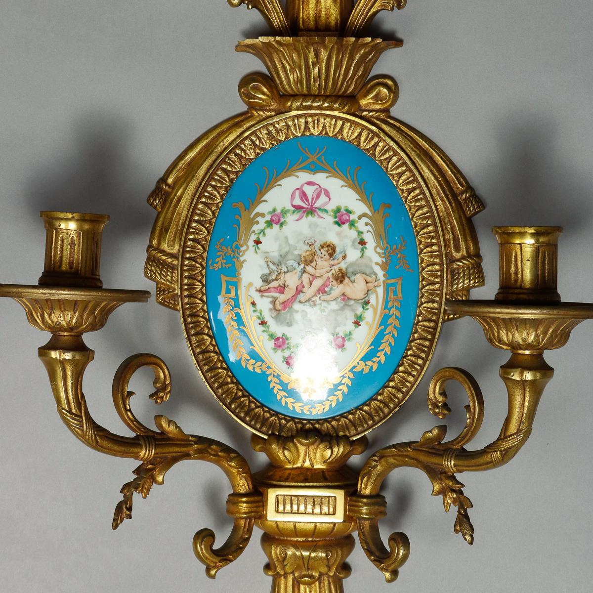 A pair of antique French wall sconces each offer a cast and gilt bronze tassel form frame housing a central hand painted and gilt Sevres porcelain plaque and having four scroll and foliate form arms terminating in candle sockets, circa