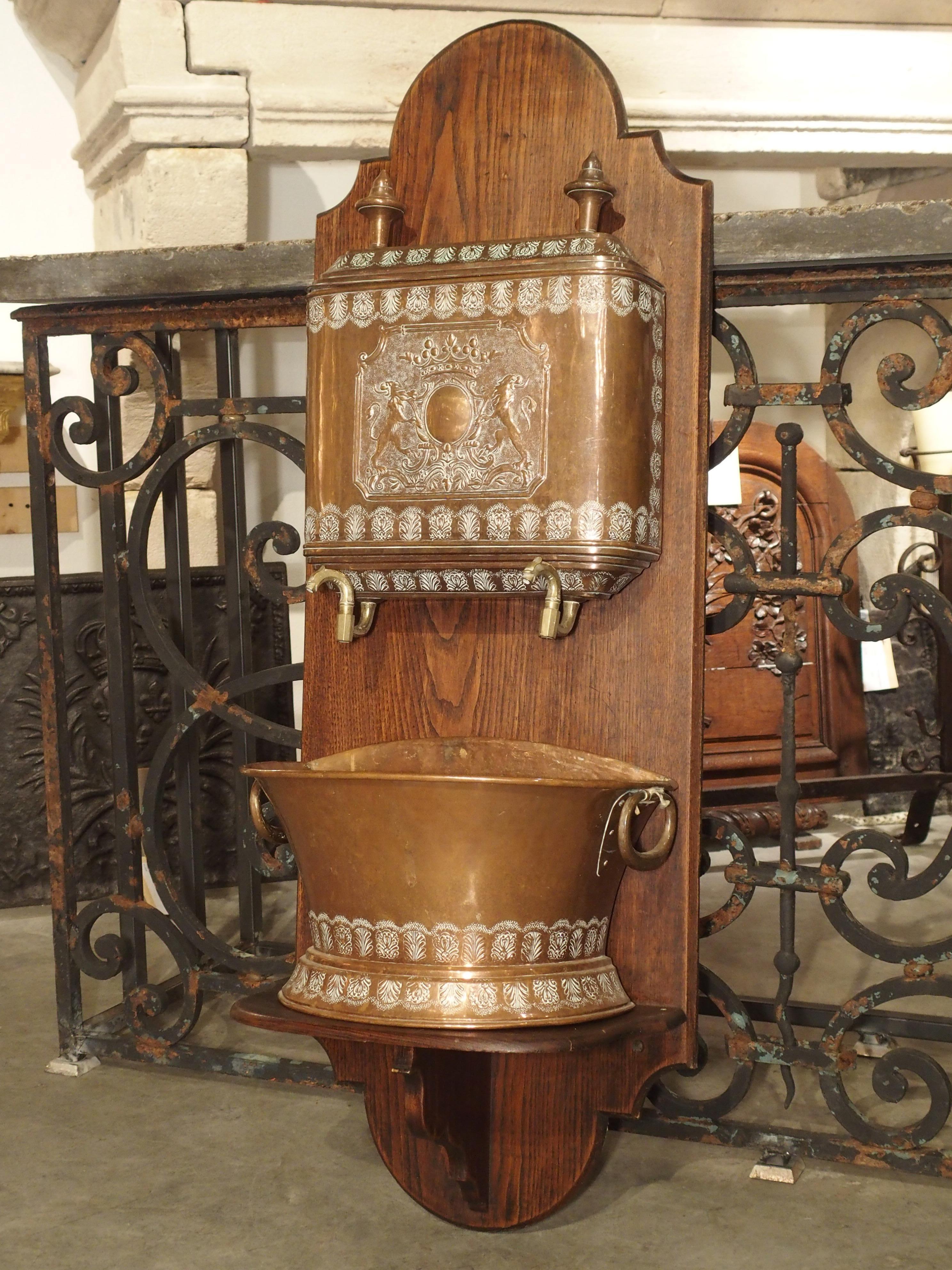 This highly worked 19th century French repousse copper lavabo on carved wooden stand has a coat of arms on the front of the upper storage tank where the water is held. Two bronze spigots would have allowed the water to empty into the bottom basin.