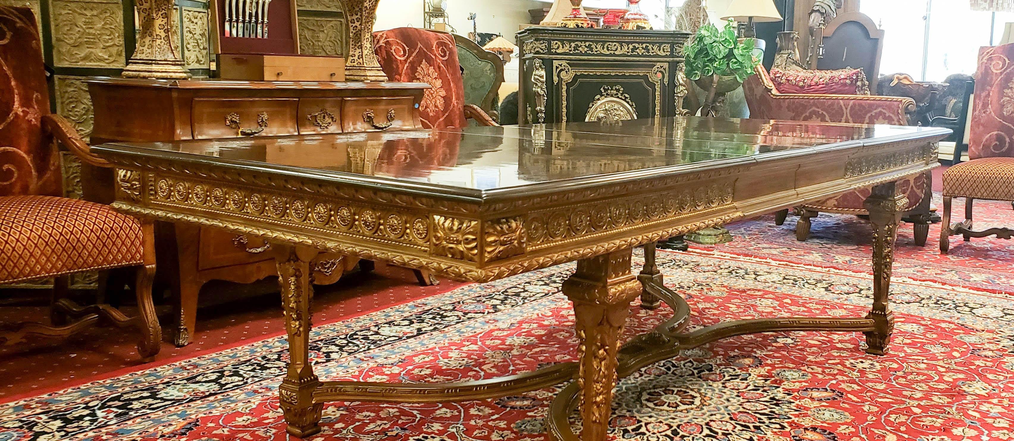 A magnificent antique French dining table. The table top is accented by a darker-tone wood border.  The apron and legs are heavily carved and finished in gilt gold, connected by a central X-shape stretcher. The three additional leaves extend the