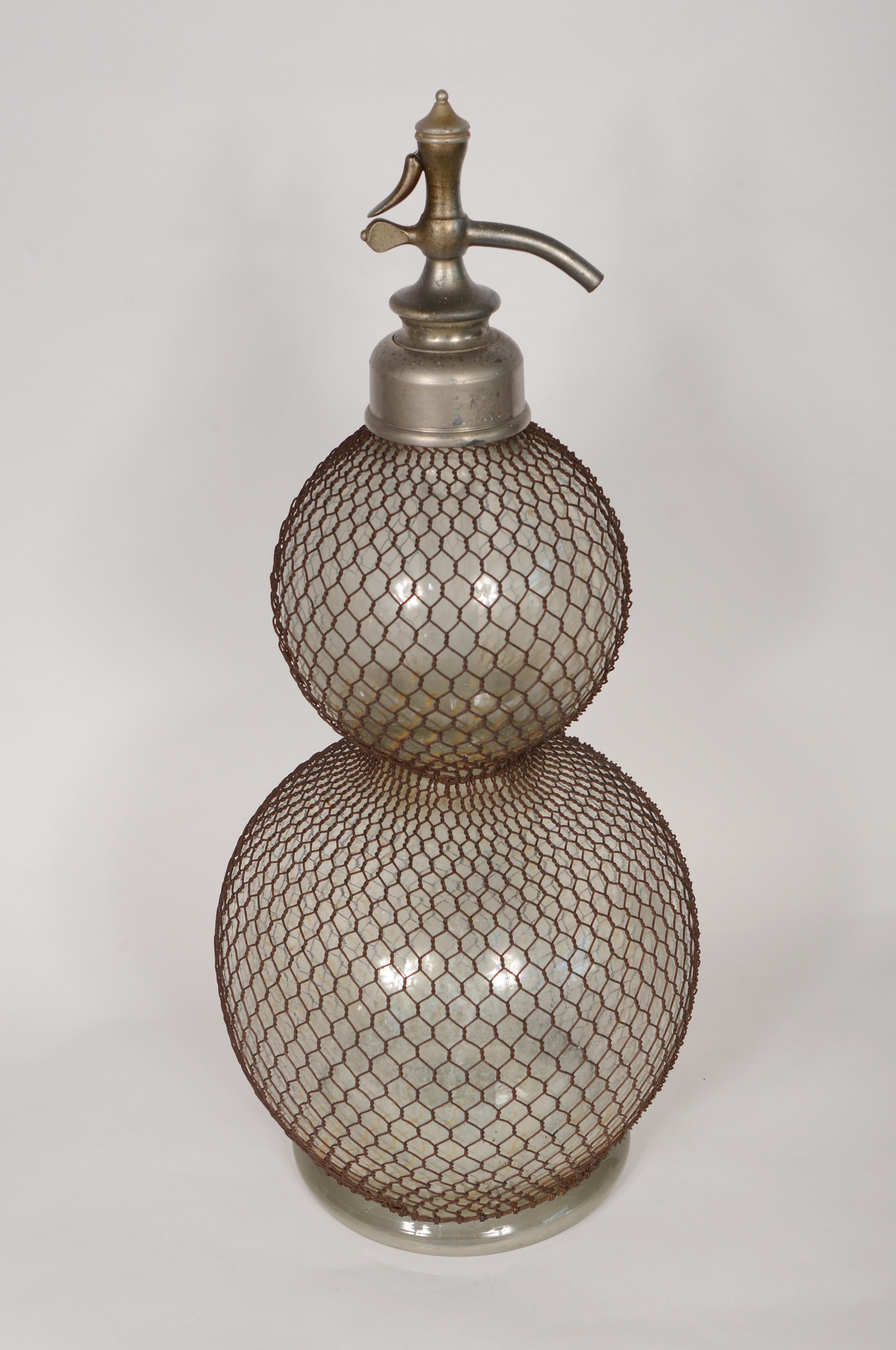 French seltzer bottle from the late 19th or early 20th century. This has wire mesh over a glass double ball form. The lever is stuck in the down position. The mesh has surface rust. There is some rust transfer to the glass. The inside of the glass
