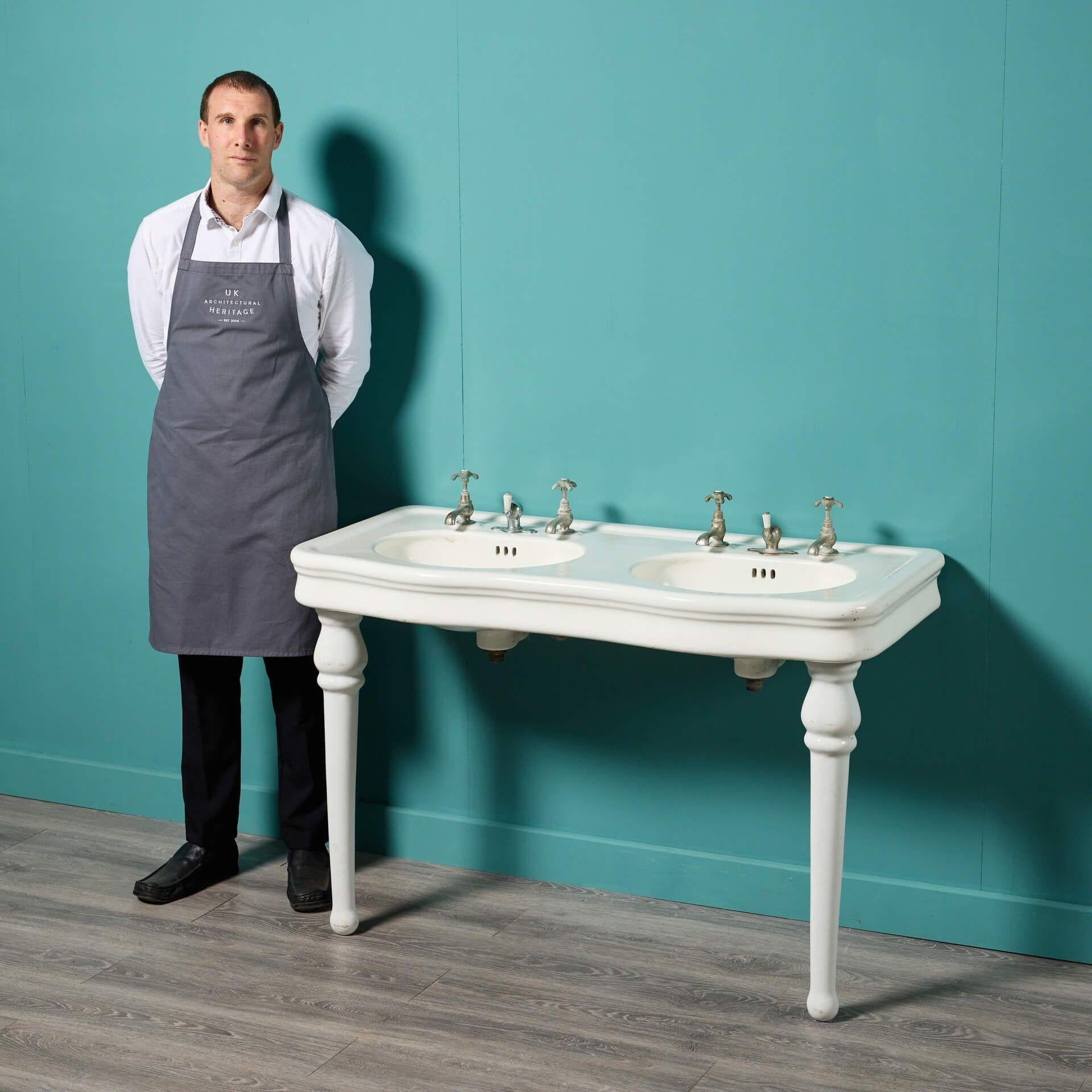 An early 20th century antique French double sink with porcelain legs. Supported by two sleek ceramic legs, this fashionable large scale sink displays elegance with its white glazed finish and curved shape; all while incorporating a range of styles