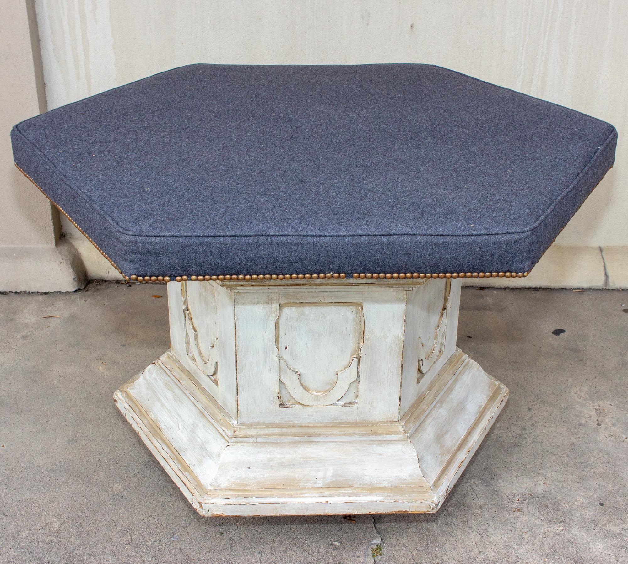 This antique French hexagonal upholstered bench or ottoman was made for a church. The carved wood base is painted white and the panels are identical on all six sides. The upholstered seat is a rich, deep gray with blue undertones, much like a slate