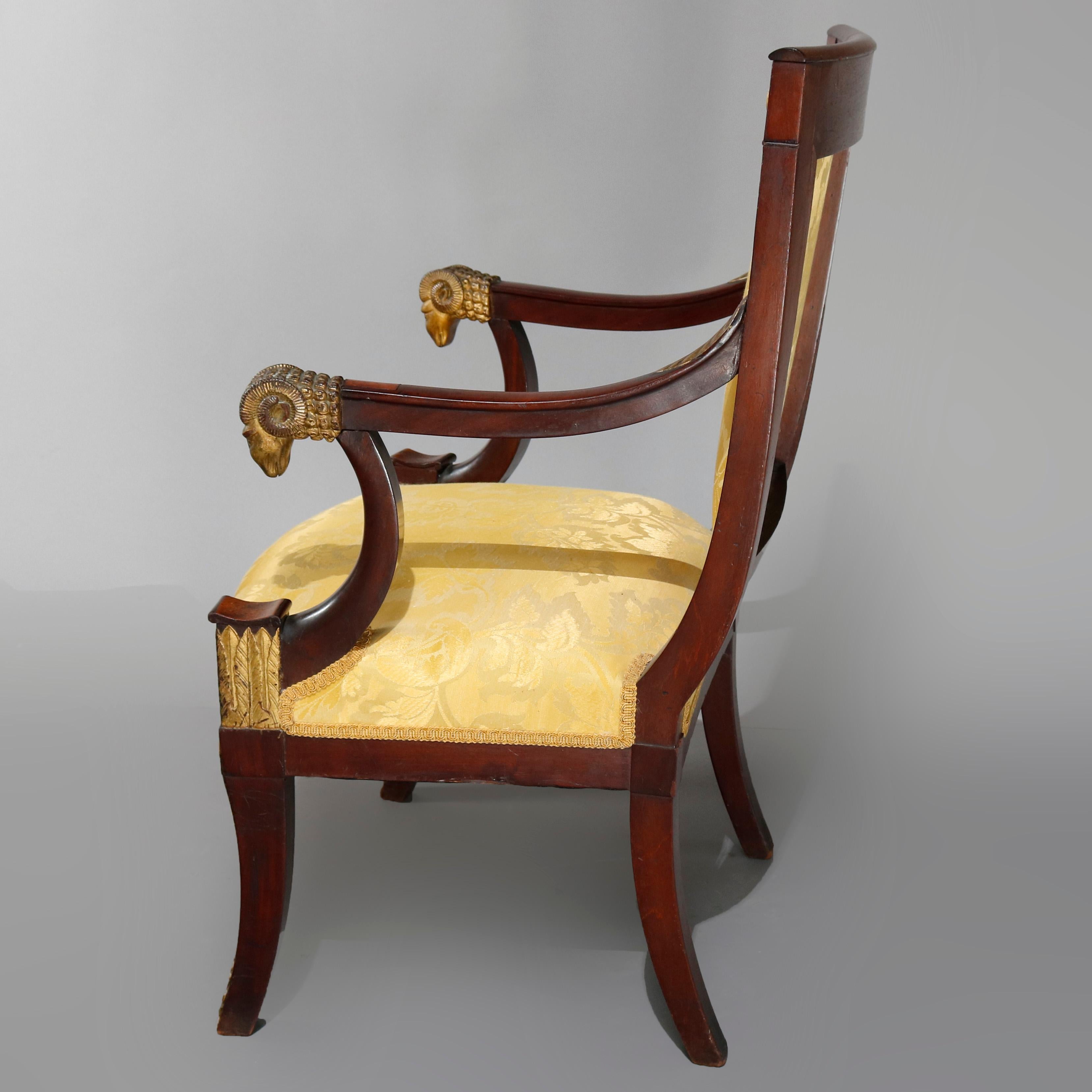 An antique early 19th century French First Empire upholstered throne chair offers mahogany frame with ormolu accoutrements including scroll, foliate and dragon crest, figural ram's head arms, Egyption reed, acanthus and palmette, circa