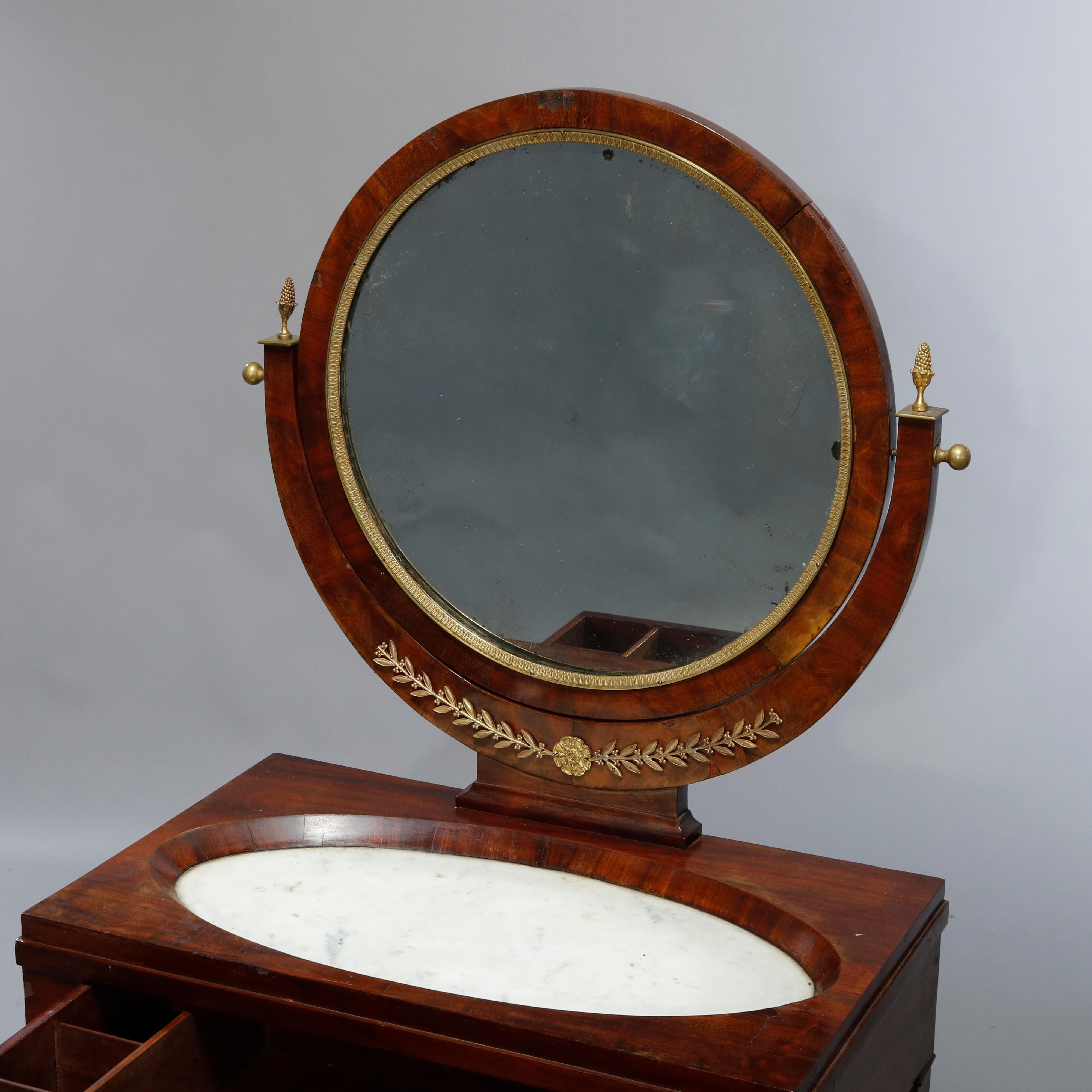 An antique French Empire dressing table offers flame mahogany construction with round swivel mirror having ormolu trim and seated in cradle having foliate ormolu mount and finals surmounting single drawer base with inset marble top and Doric column