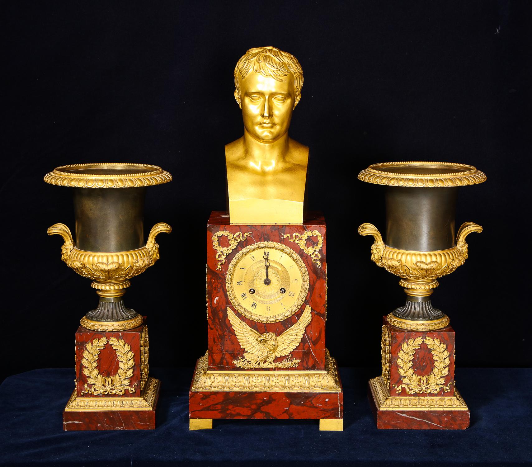 A superb antique French Empire gilt bronze, patinated bronze and carved rouge marble three piece figural clock garniture set of exquisite craftsmanship. The unique clock is embellished with a fine gilt bronze bust of Napoleon on a rouge marble base