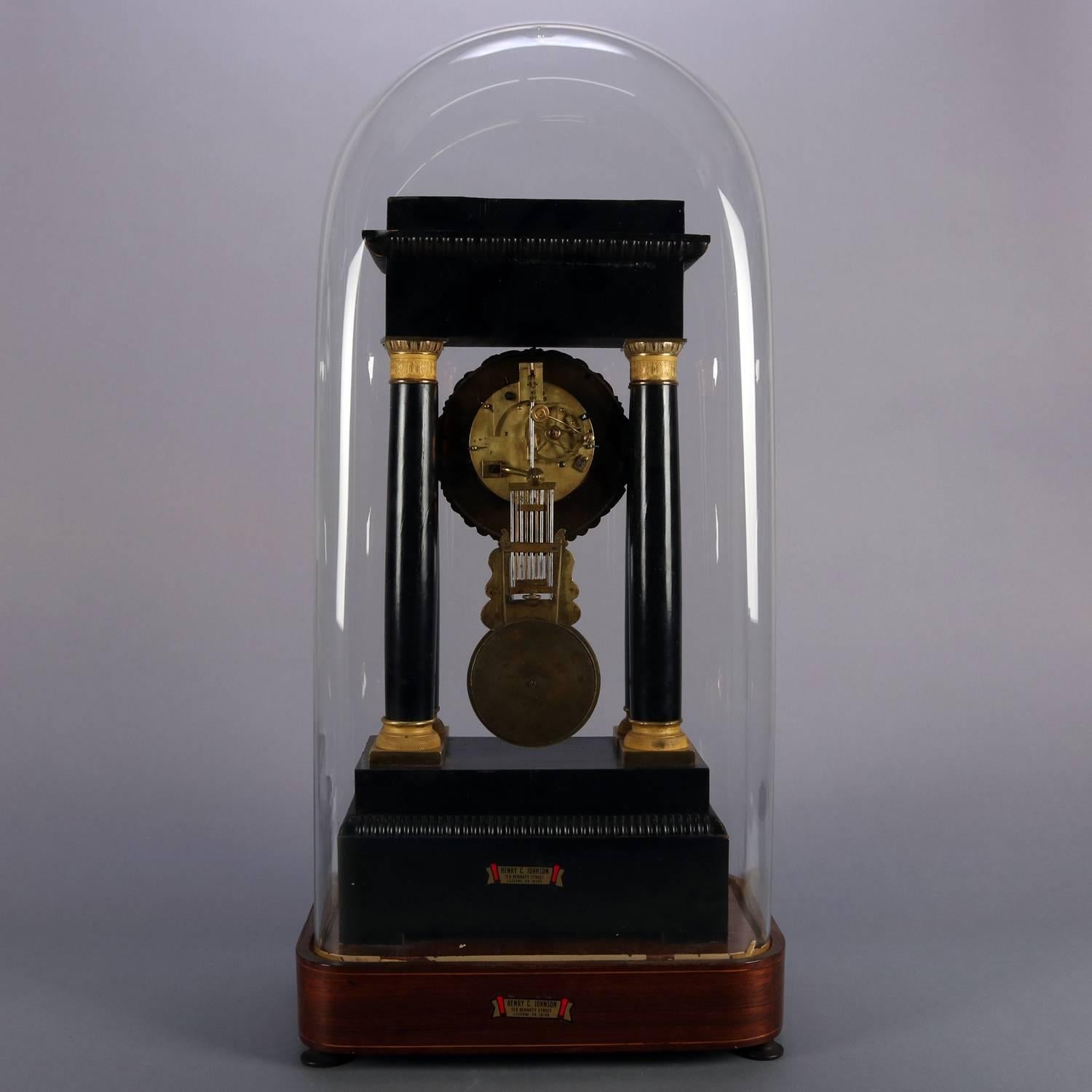 Antique mahogany French Empire portico clock features ebonized and bronze case with mahogany marquetry of satinwood foliate inlay and banding, bronze column capitals and bases, machined dial with Roman numerals, bronze mask pendulum, works signed