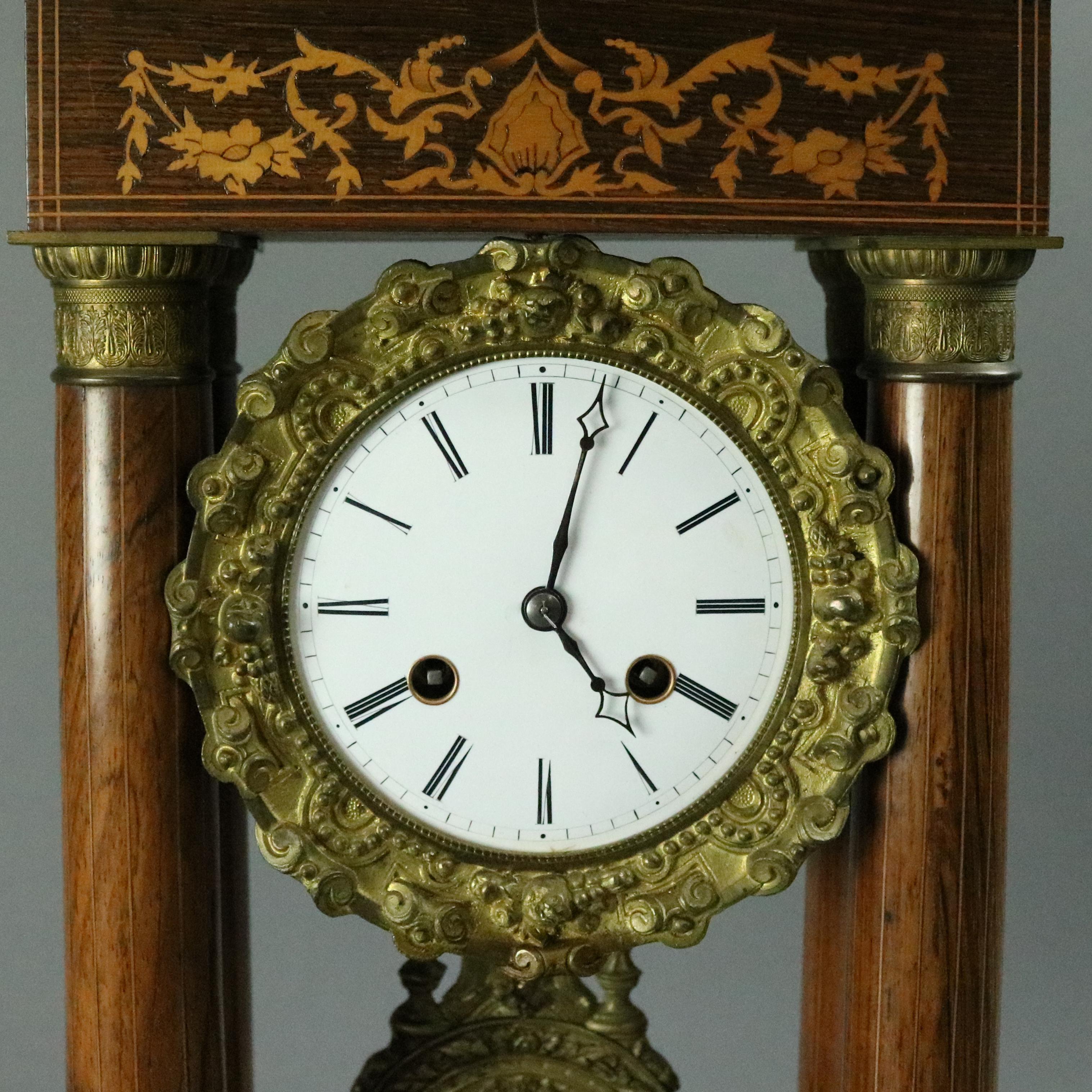 Antique mahogany French Empire portico clock features mahogany marquetry and bronze case with satinwood foliate inlay and banding, bronze column capitals and bases, porcelain dial with bronze surround, bronze mask pendulum, works signed Medaille