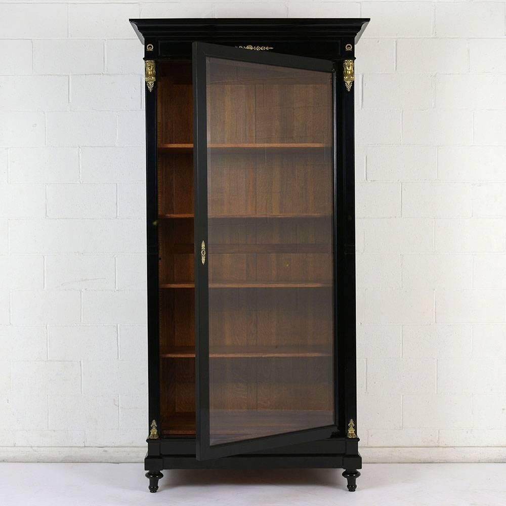 This 1900s French Empire-style bookcase is made of mahogany wood with an ebonized and lacquered finish. The Classic design features a top cornice and decorative legs with decorative brass accents. The single beveled glass door bookcase features