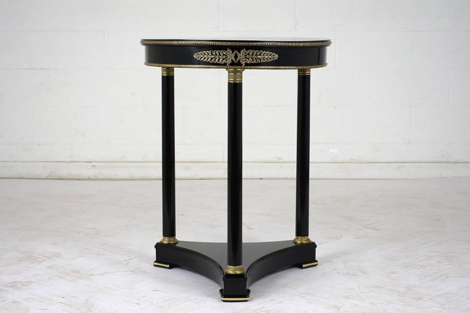 This 1900s French Empire-style round side table is made of mahogany wood with an ebonized and lacquered finish. The table has a triangular pedestal base with simple columns. The table is accented by bronze decorative bands and ormolu accents. This