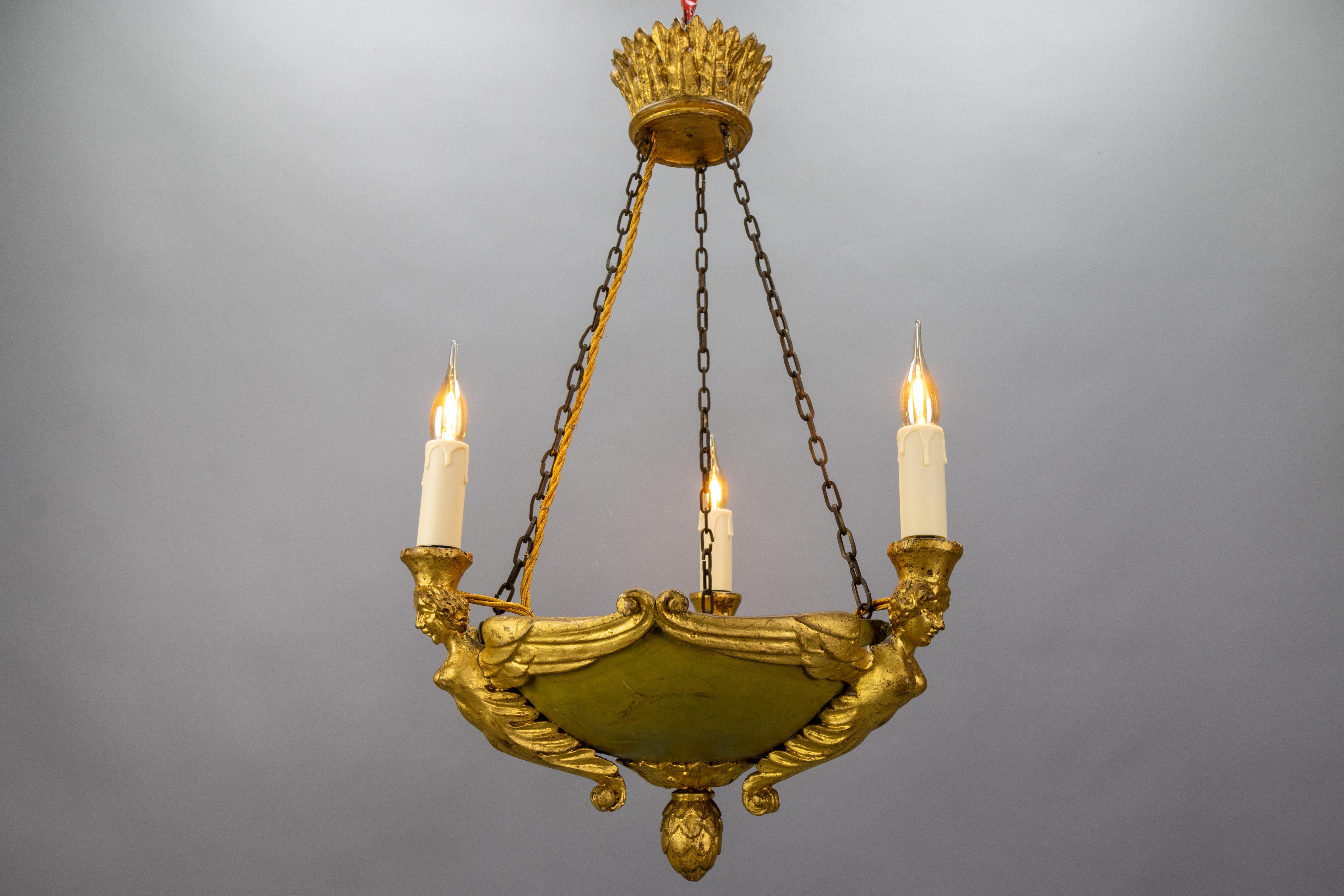 Antique mid-19th-century French Empire-style gilt linden wood chandelier featuring three winged female figures, an olive-green painted bowl, and an ornate crown canopy. Recently rewired with new E14 sockets. Elegant neoclassical design, rich