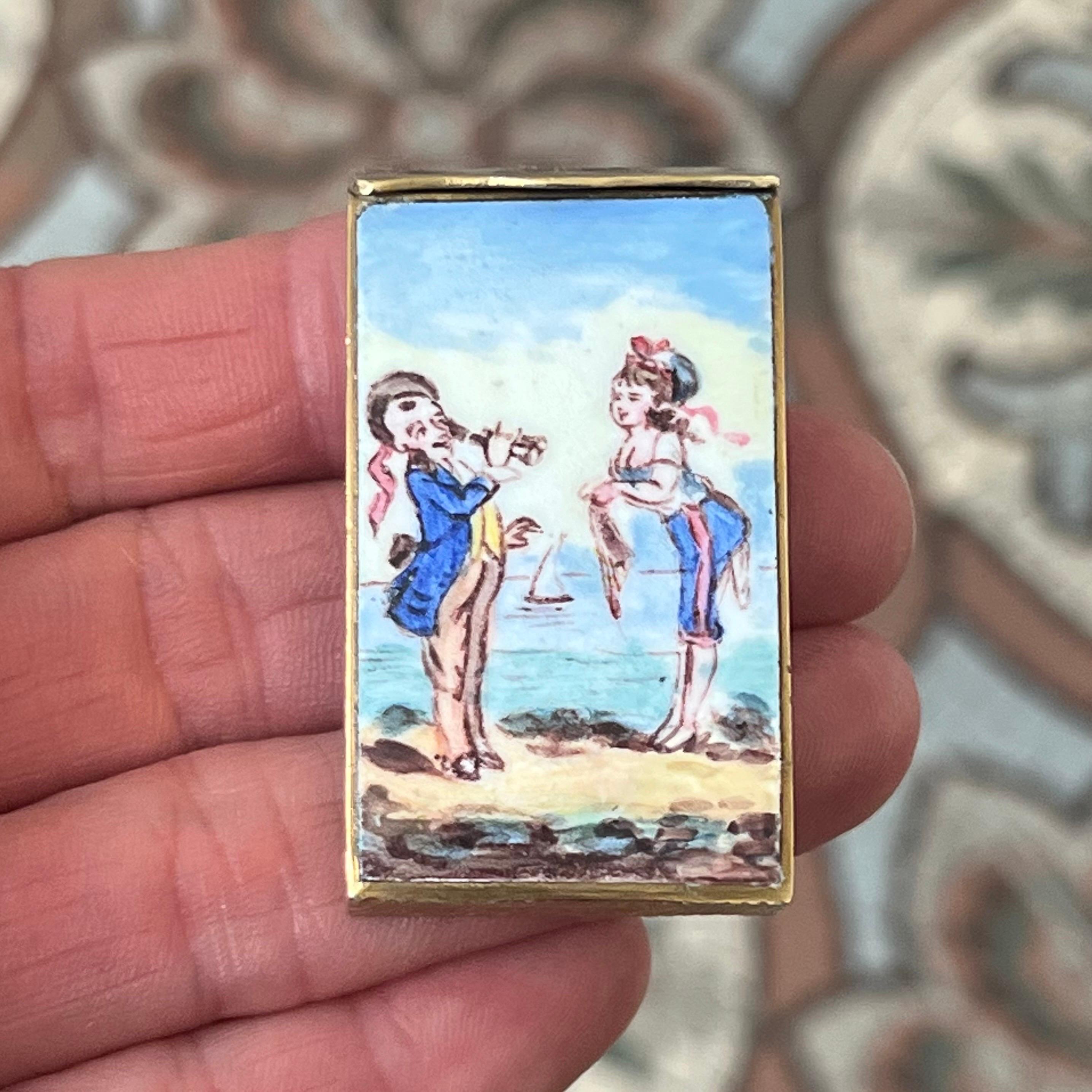 A delightful antique French vesta case (match safe), finely crafted in gilt brass and decorated with a colorful hand-painted enamel plaque. The enamel scene depicts a playful seaside motif: a young sailor serenading a girl with a flute, while a