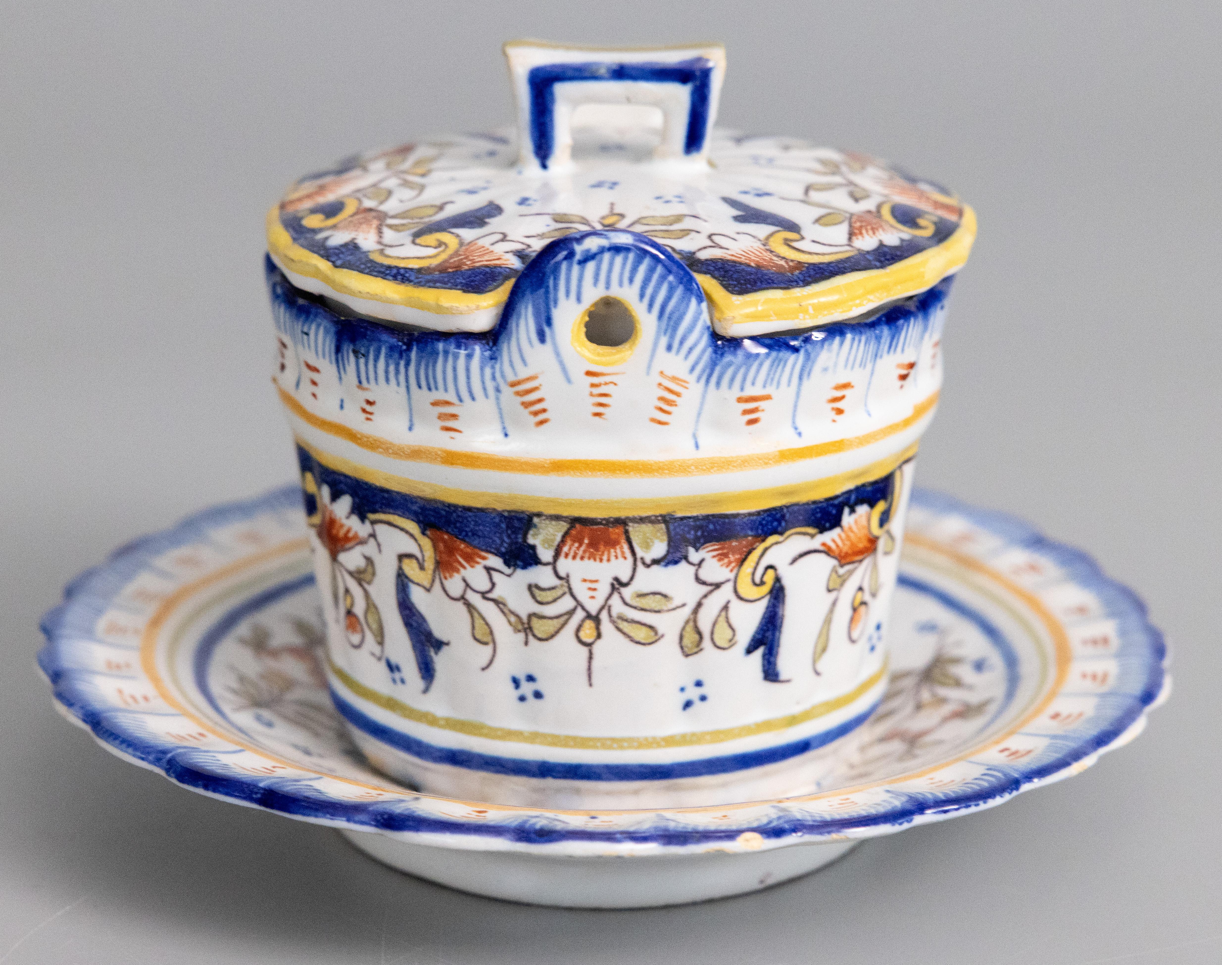 A superb antique French faience Desvres lidded butter bowl dish tureen with attached underplate, circa 1900. Maker's mark on reverse. This charming dish has lovely scalloped edges, a hand painted armorial crest, and floral decoration in the