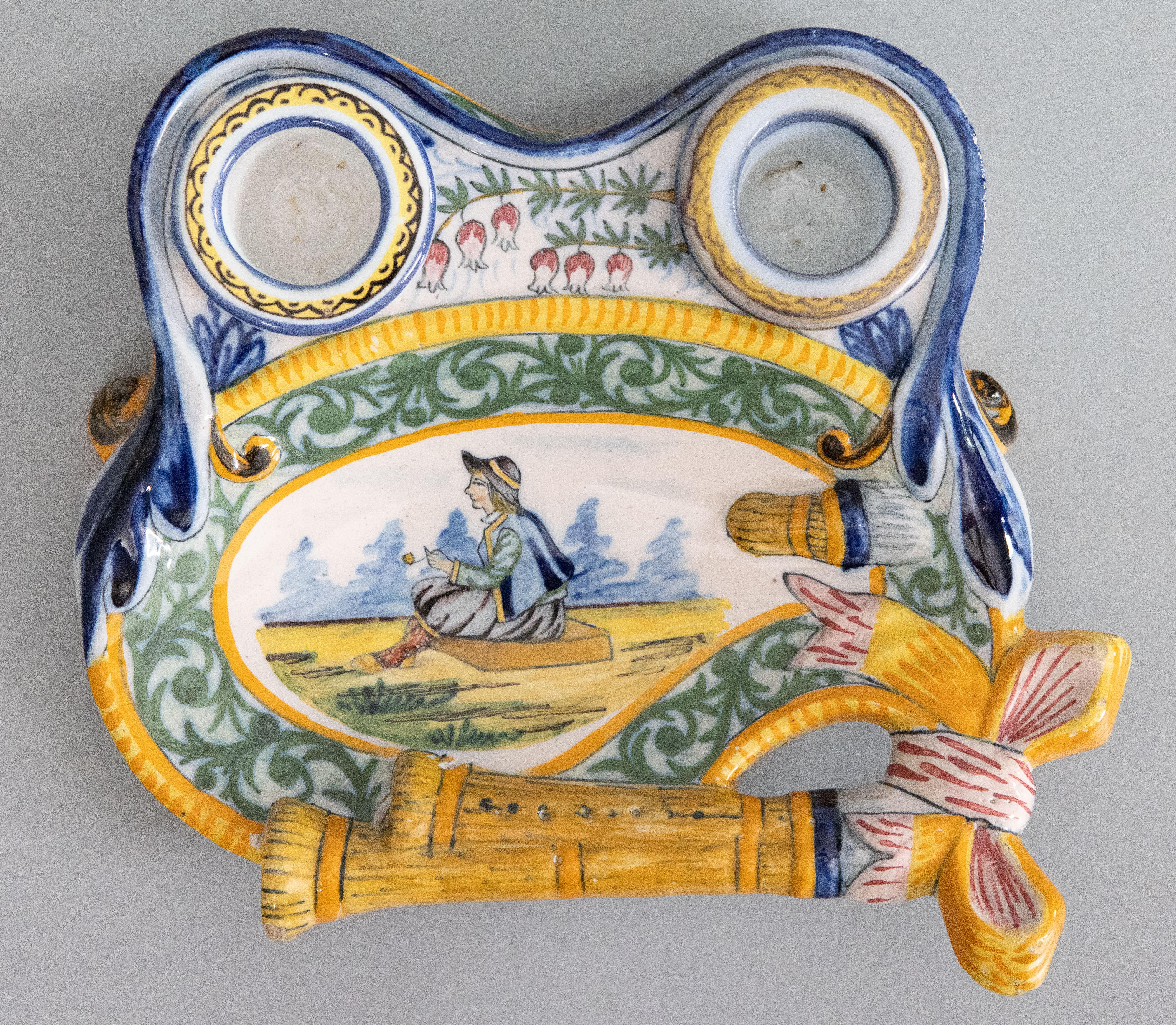 A charming antique French faience Quimper double inkwell and pen tray desk set, circa 1900. This gorgeous inkwell is hand painted with a traditional Breton man surrounded by bagpipes, green foliate scrollwork, and beautiful flowers. It would be a