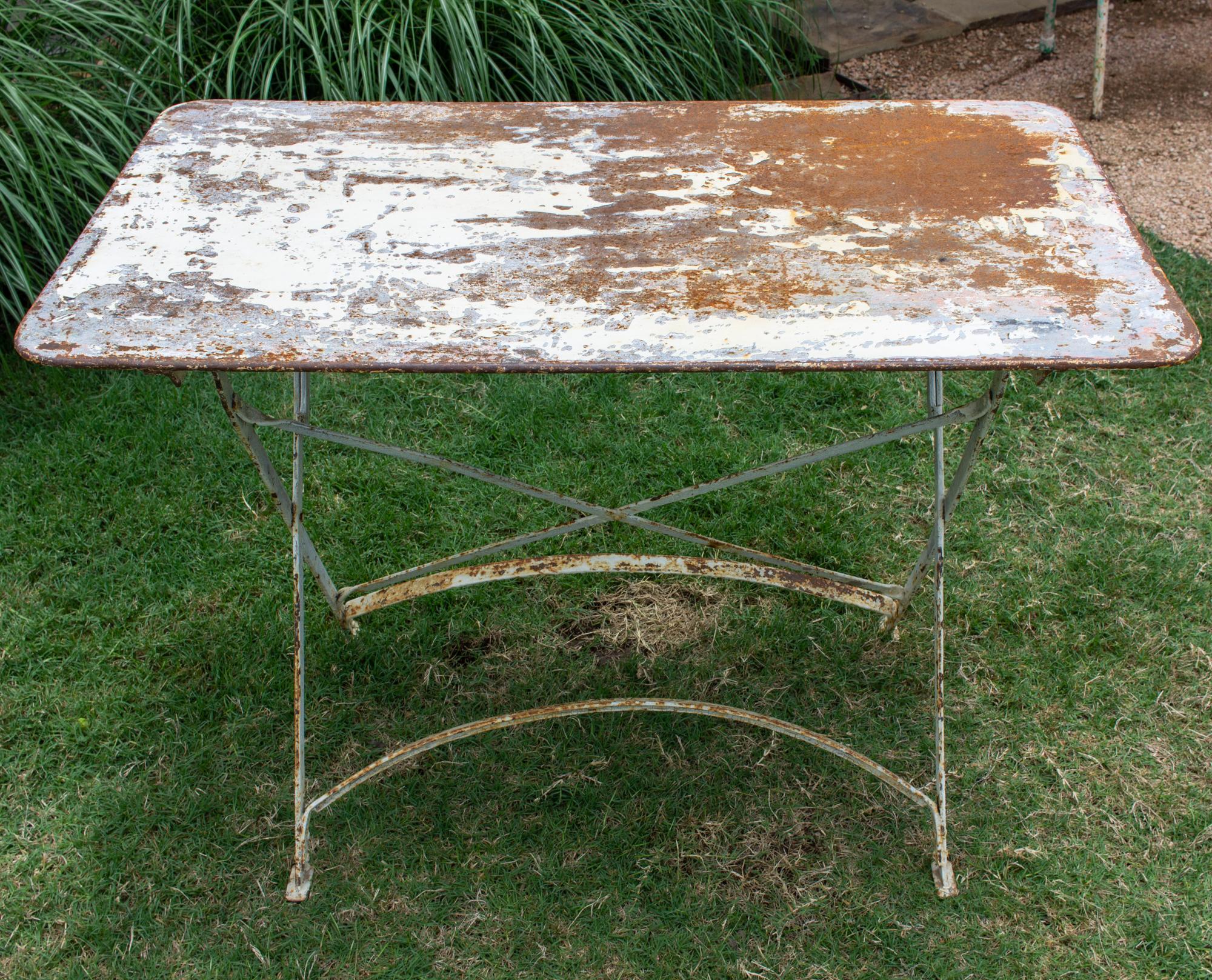 This antique French garden table is crafted in metal with a distressed painted finish. The table folds flat for storage when not in use. The top has rounded corners and a wonderful patinated finish. The base has an X detail on one side and curved