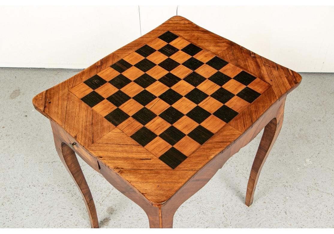 A small parquetry walnut table with inlaid chess board top in lighter and black wood squares. Two small frieze drawers for two sets of Italian made chess pieces, in silver tone and gilt tone metal. With a serpentine skirt and raised on tall curved