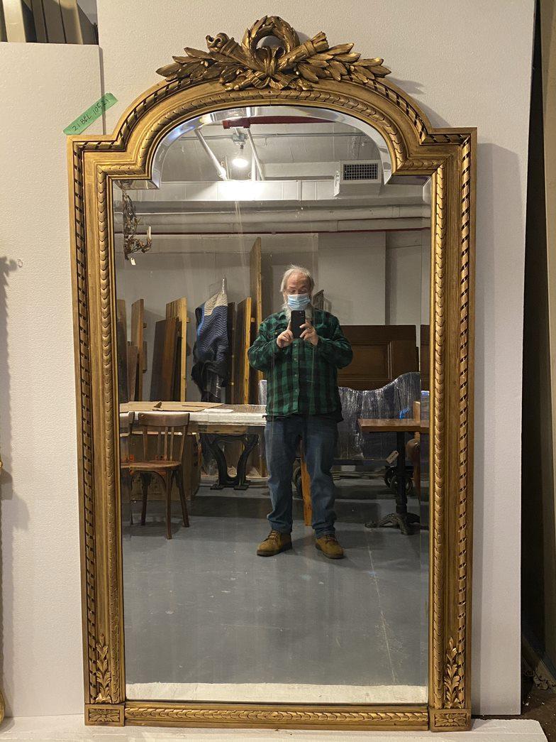 Large French gold carved wood frame beveled mirror with decorative details and a torch motif at the top from the early 20th Century. Imported from Europe. Original distressing on mirror. This can be seen at our 333 West 52nd St location in the