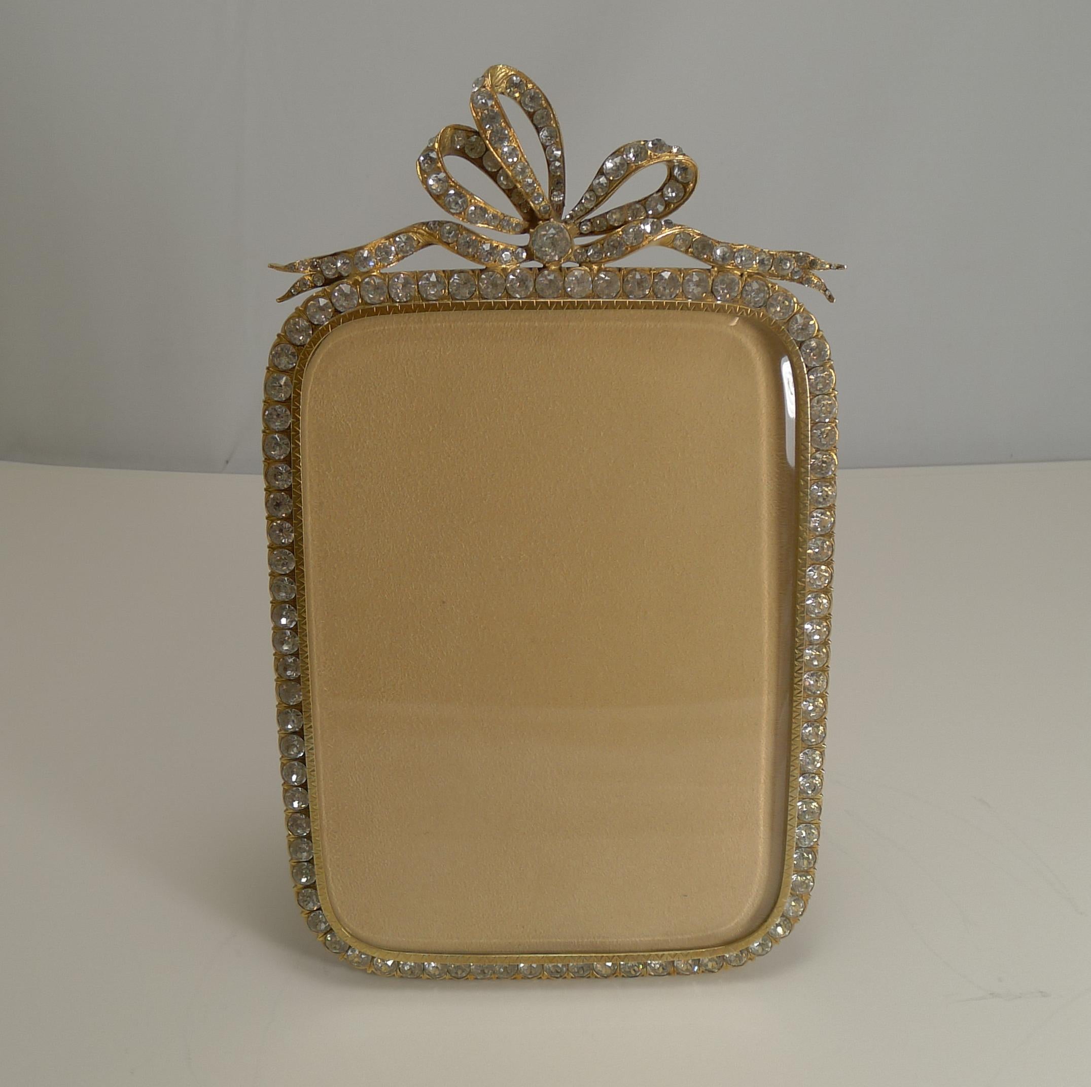 A rare and fabulous large photograph frame made from gilded bronze, a good solid frame smothered in gold and inset with sparkling soft paste stones creating a piece of Hollywood glamour.

The top is crowned by a singe large tied ribbon and bow,