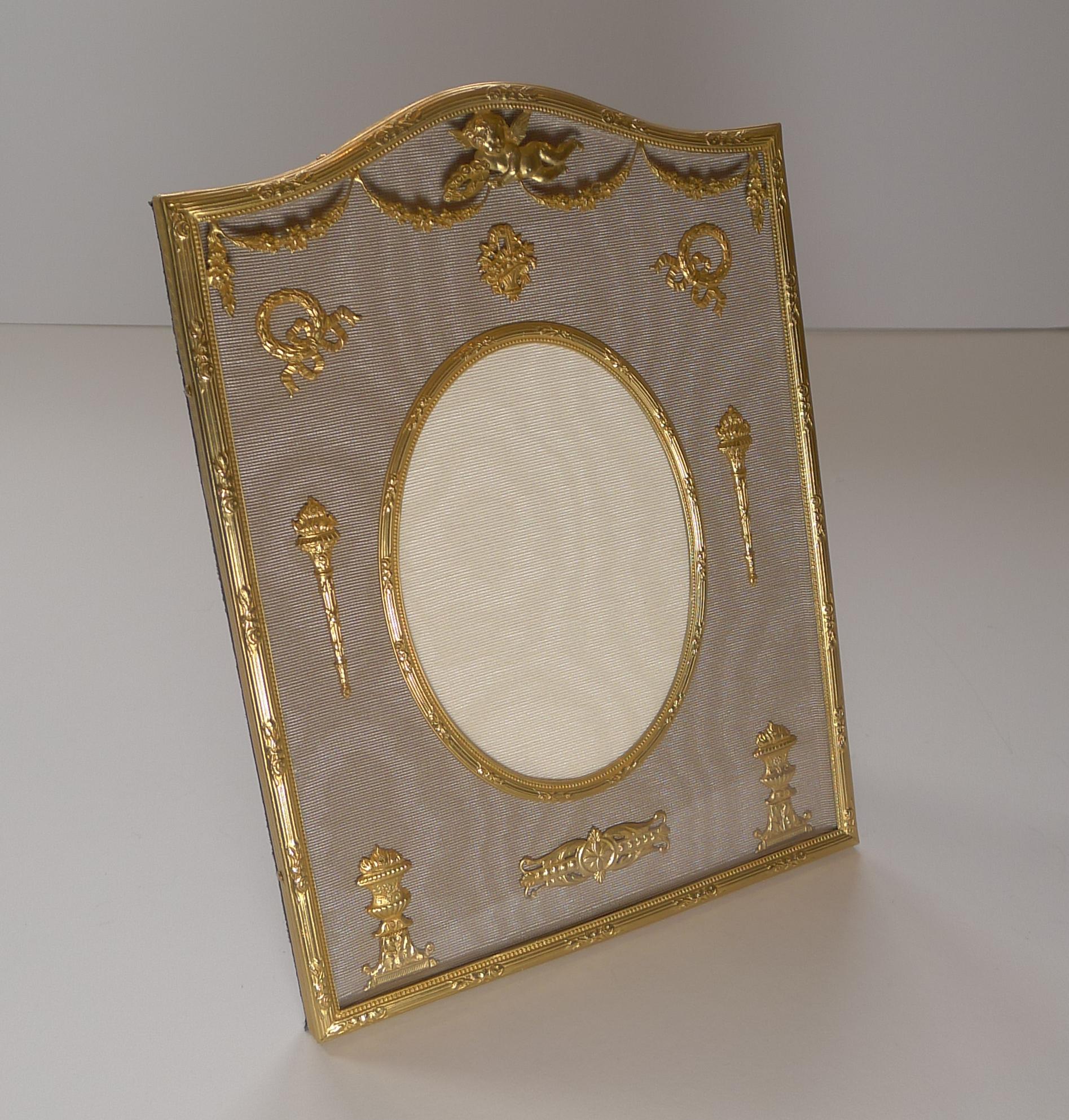 A handsome bronze dore / Ormolu photograph frame with an arched top and a central oval aperture behind the glass.

The taupe coloured taffeta is mounted with gilded bronze motifs and to the top there is a mount on top of the glass with a central
