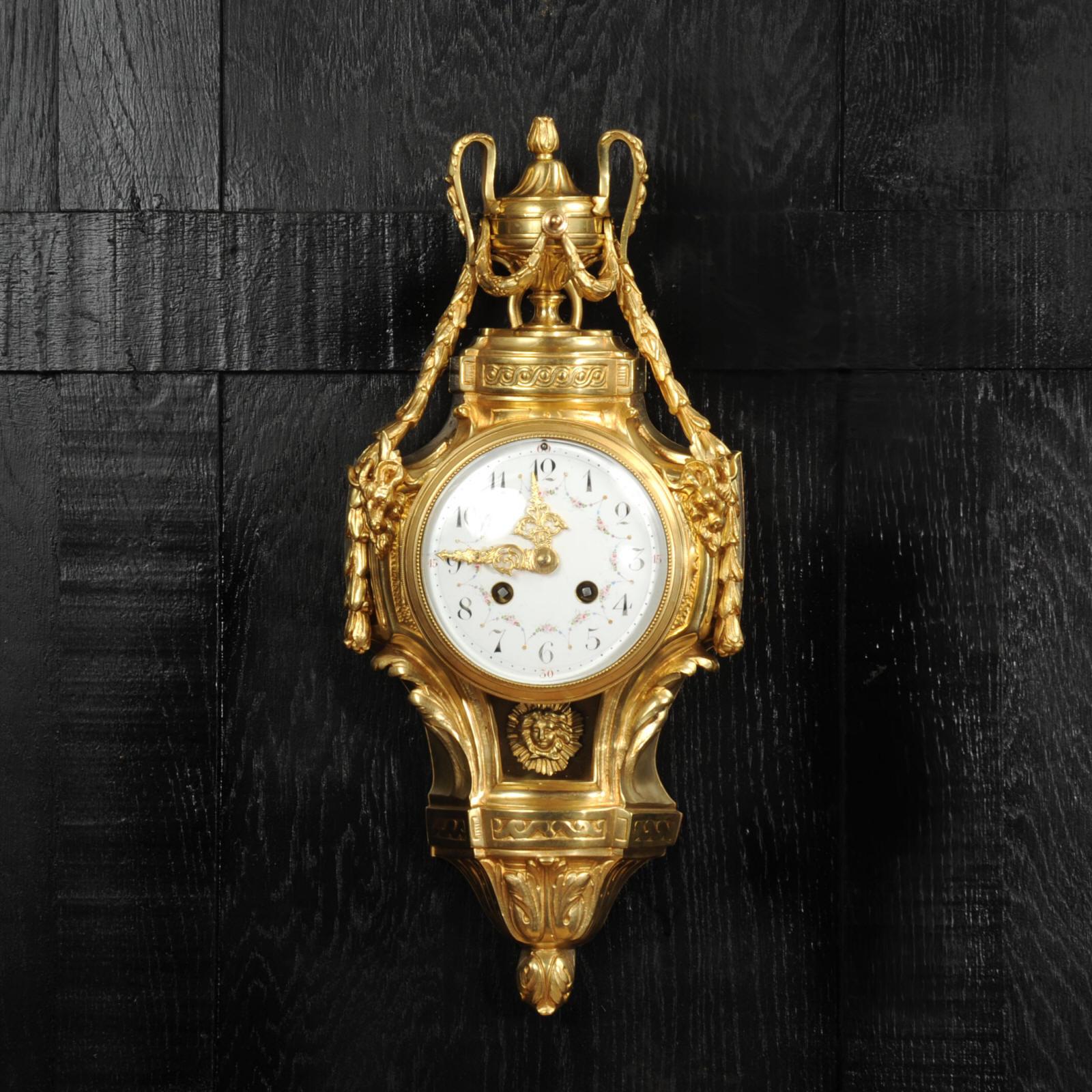 Precioso reloj de pared de cartel clásico de estilo Luis XVI, magníficamente realizado en bronce dorado. De elegante diseño, con guirnaldas de laurel drapeadas, cabezas de carnero y una abertura acristalada bajo la esfera que permite ver oscilar
