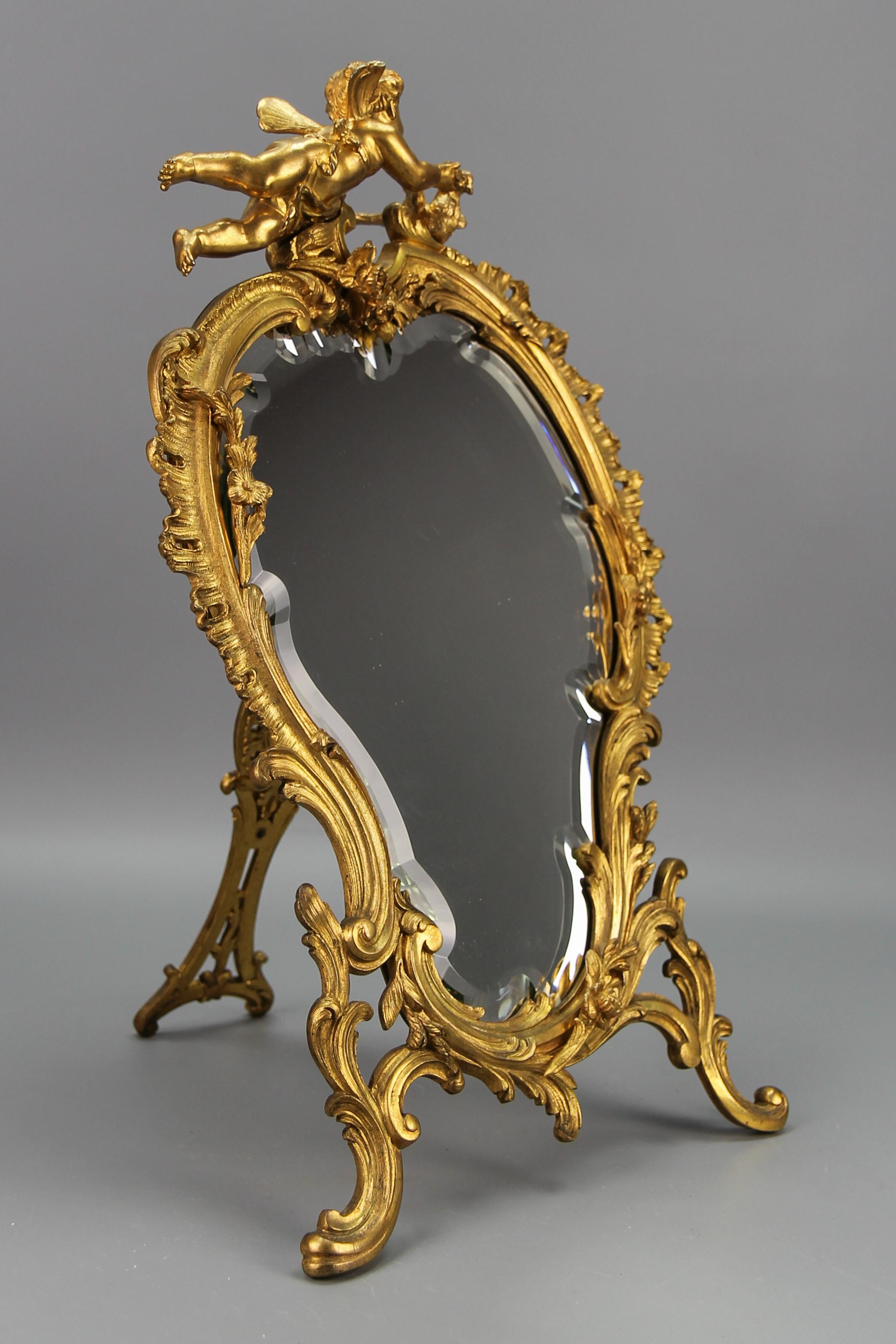 Antique French gilt bronze Rococo style desktop mirror with cherub and bird from the early 20th century.
Adorned with typical Rococo or Louis XV-style scroll, floral, and foliate motifs around the beautifully shaped and faceted mirror, this large