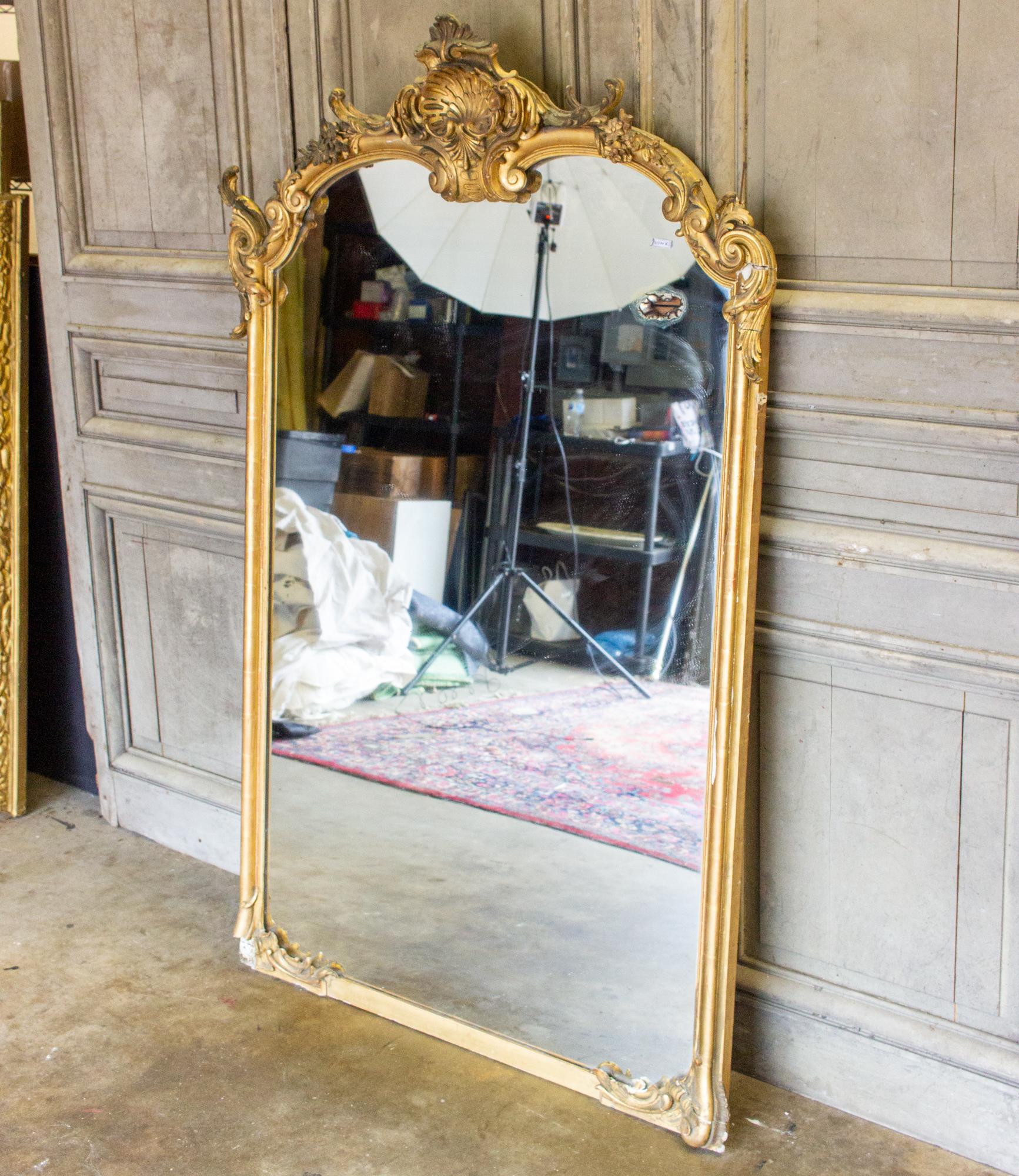 This beautifully decorated French gilt full-length mirror features a large and ornate shell cartouche at the top as well as floral carvings and scroll work at each side, corner and surrounding the cartouche. The mirror is in good shape, with one