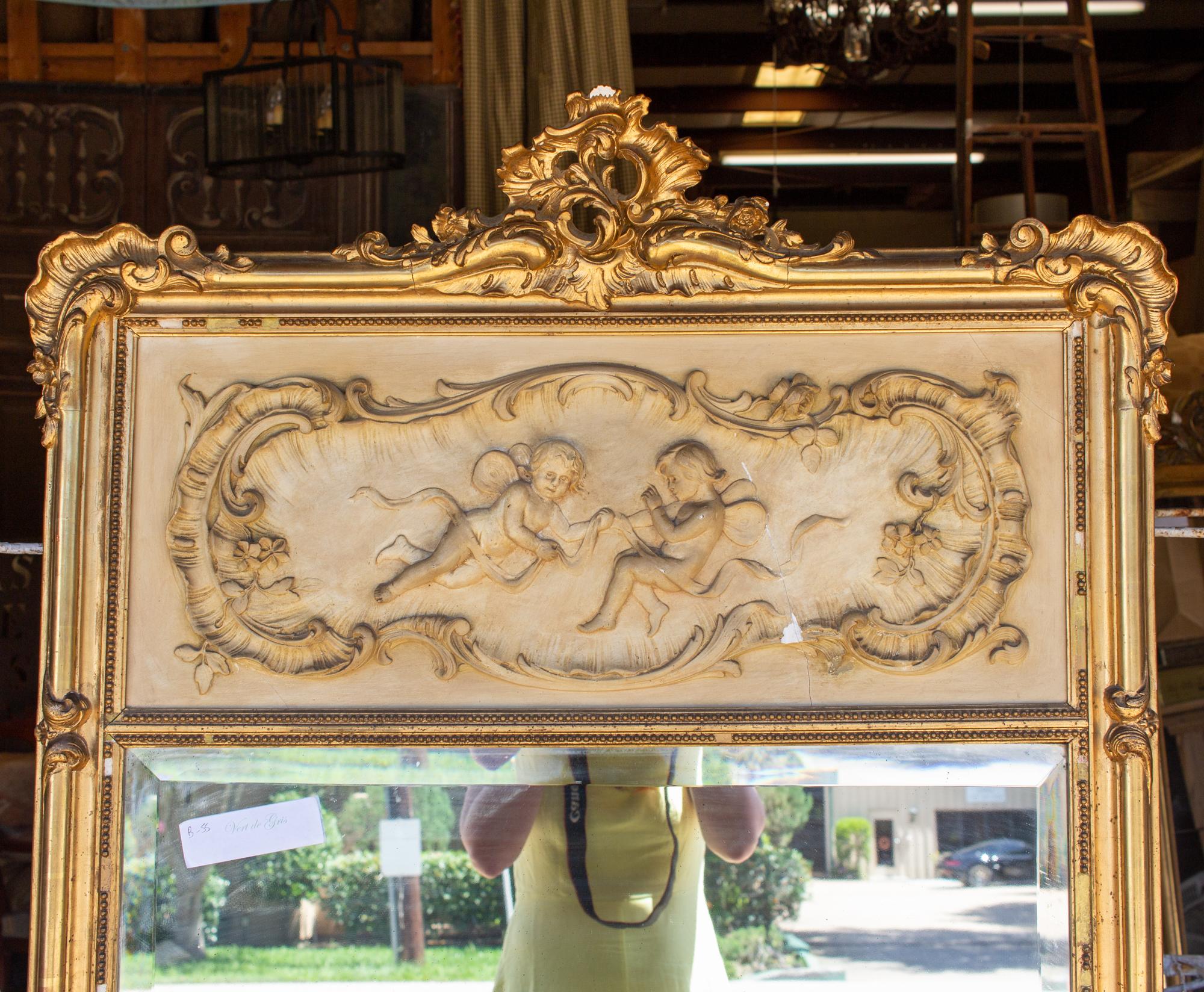 This antique French gilt trumeau mirror has a wonderful plaster panel inset at the top, with two fairy-like figures, scrollwork and floral accents. The mirrored portion features a beveled edge and the frame has decorative accents at the corners and