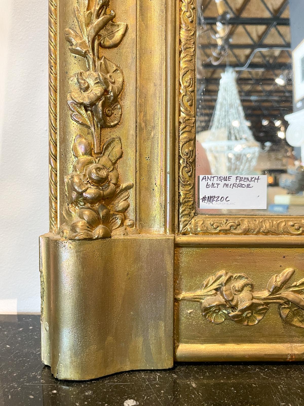 This antique French gilt wall mirror is rectangular in design with floral accents around the frame which has a heavier width at the bottom. The original, antique glass is showing signs of age, with the crystalline structure most visible around the