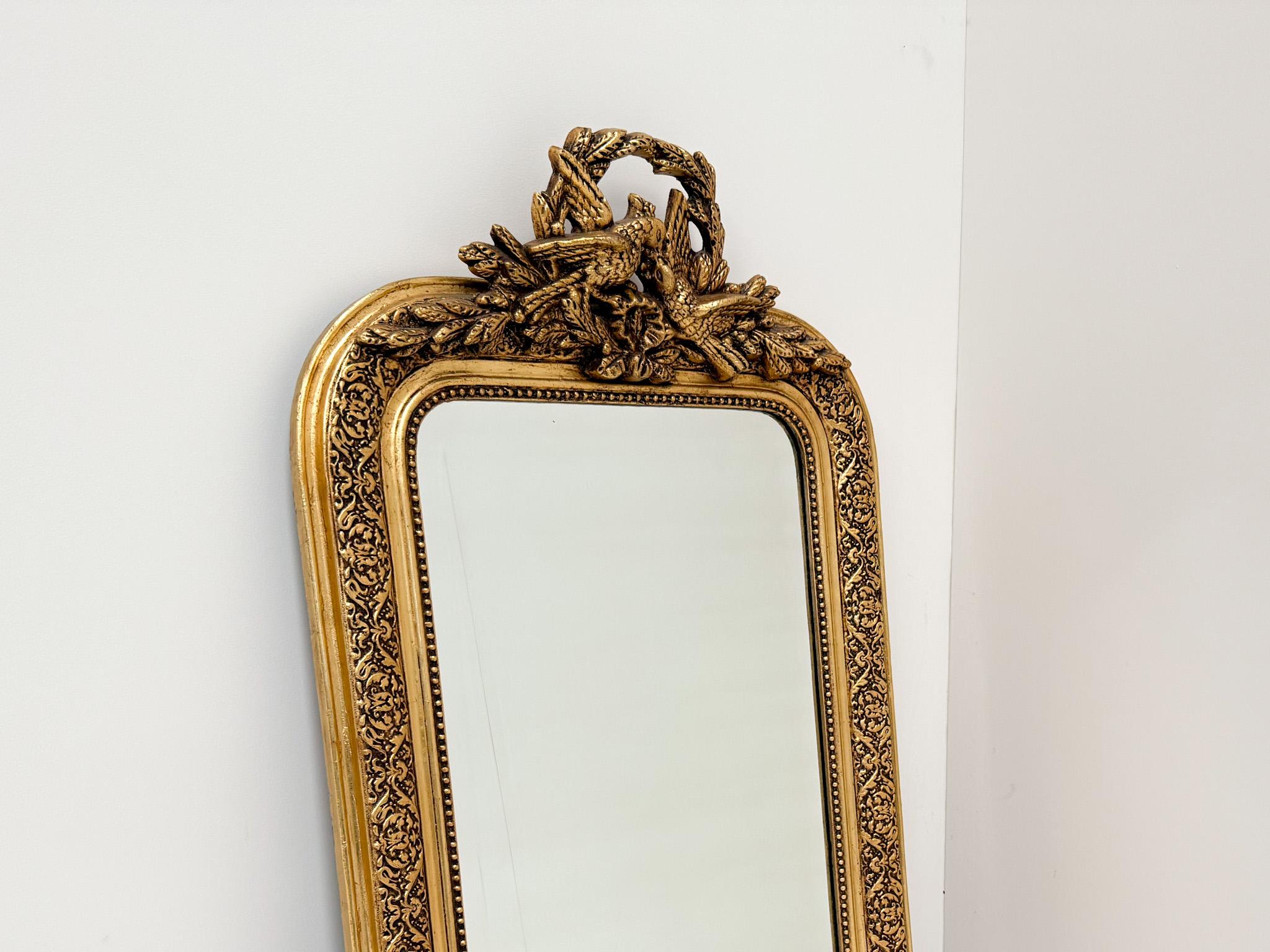 Bellissimo specchio francese in legno dorato risalente al XIX secolo. Lo specchio presenta una cornice riccamente decorata con intricati motivi floreali e uno splendido stemma ornato da uccelli, foglie di alloro e nastri. La finitura in legno dorato