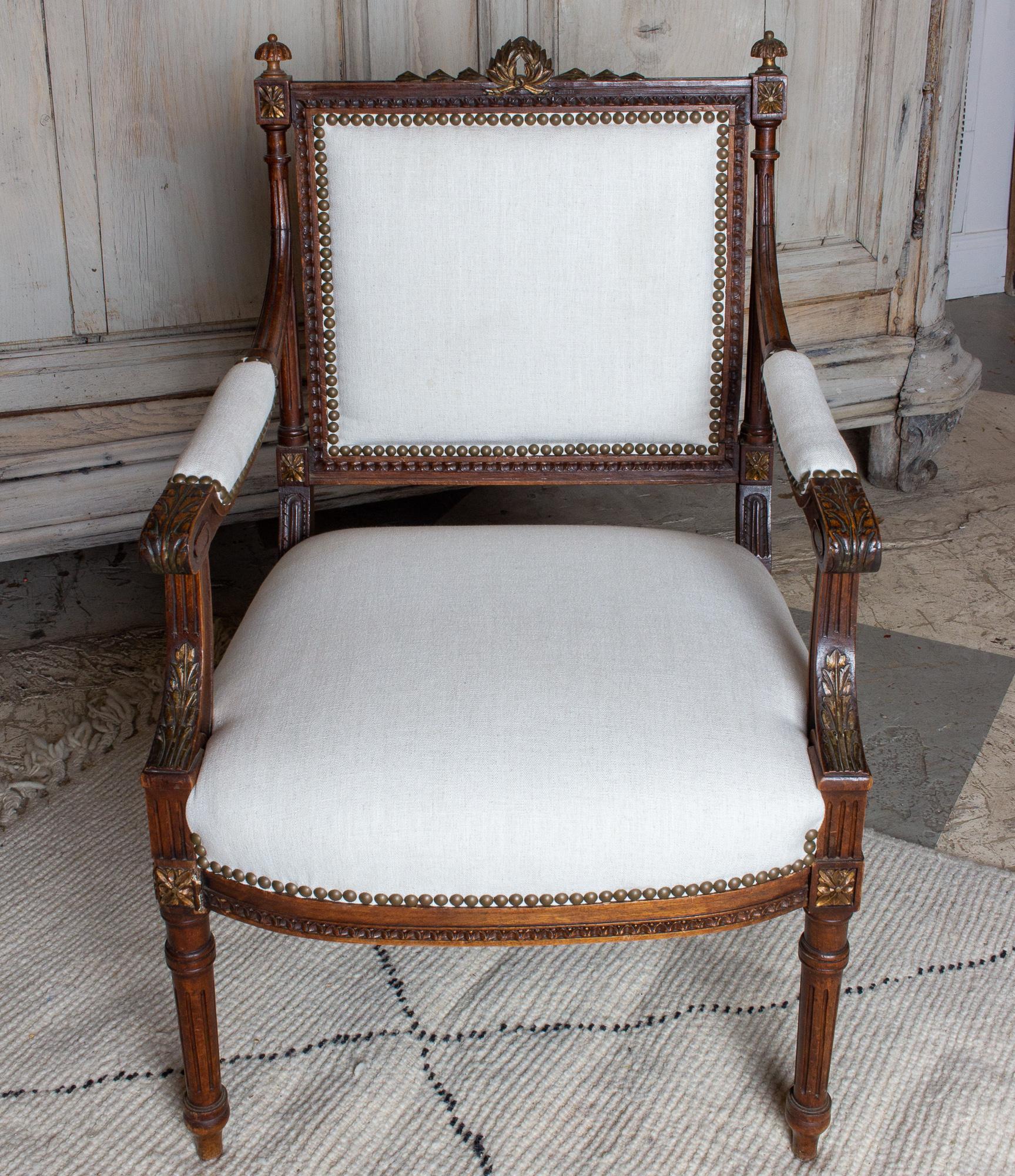 A lovely pairing of antique French hand-carved Louis XVI style armchairs with new linen upholstery. The chairs have classical carvings and gilt accents. Antiqued brass nailhead trim and a neutral linen fabric ensure both elegant and versatile use in