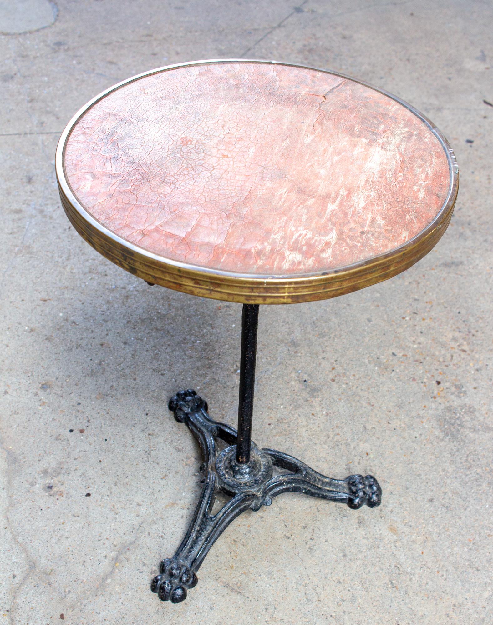A classic French iron bistro table, this piece features a black iron base with paw-foot detail and a brass rim surrounding a distressed leather top. The leather is worn and cracked, and is a rich cognac color that leans red. The top and brass edge
