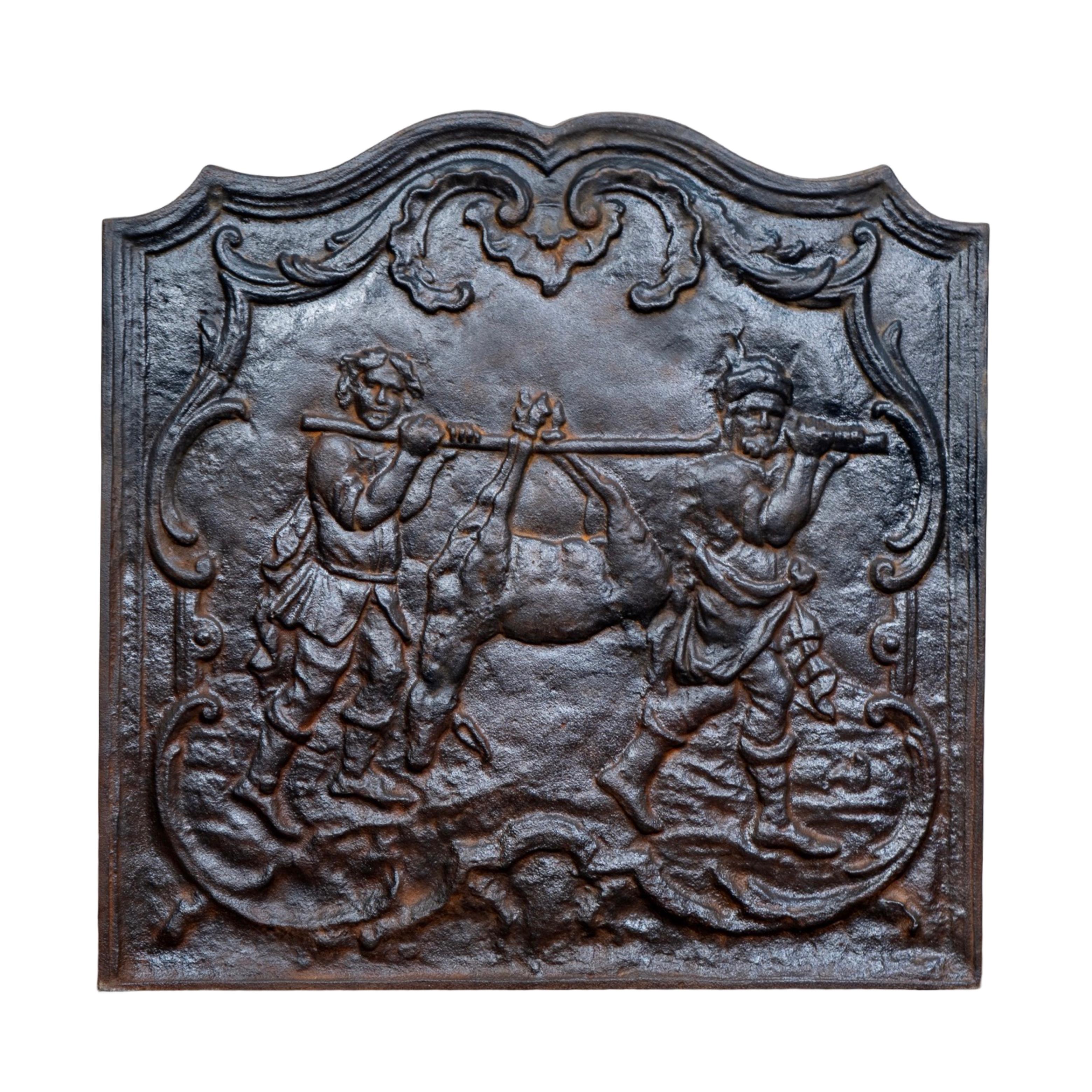 A richly detailed 18th-century French cast iron fireback featuring a vivid hunting scene in high relief. The central motif portrays two figures carrying a game animal suspended on a pole—an iconic depiction often associated with harvest or biblical