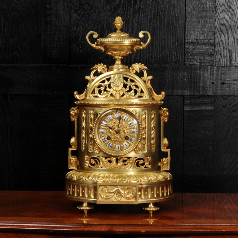A beautiful original antique French clock. It is classical in the Louis XVI style, an oval shaped body pieced with scrolling acanthus, stylised strap handles with rams heads and an urn with pineapple finial. The pendulum can be seen gently swinging