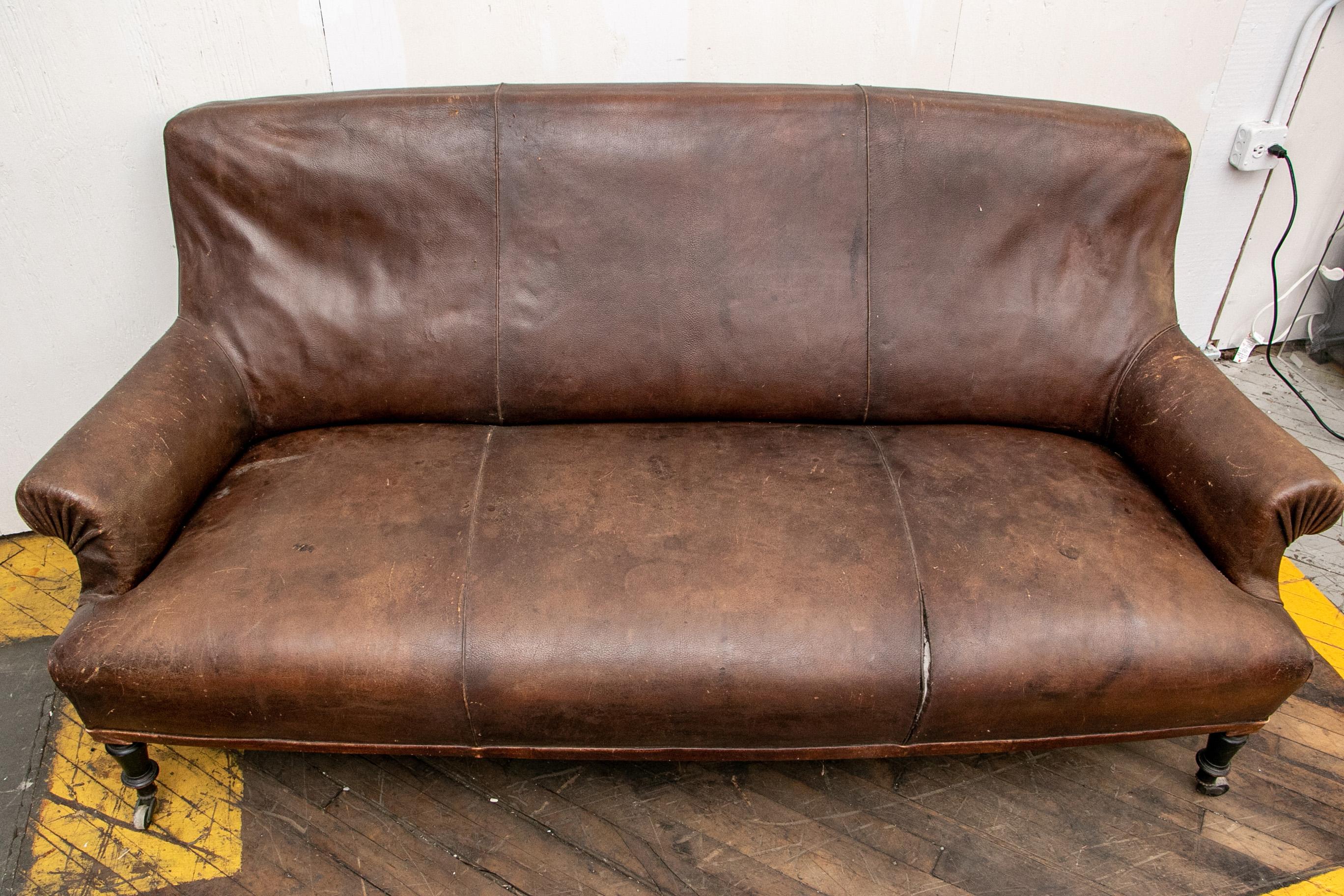 A three-seat sofa in dark brown leather, with pleated scrolled arms. Dark stained turned front legs and curved back ones, all on casters. 

Condition: expected overall wear due to age and use, split to a seat seam, scrapes to the legs however the