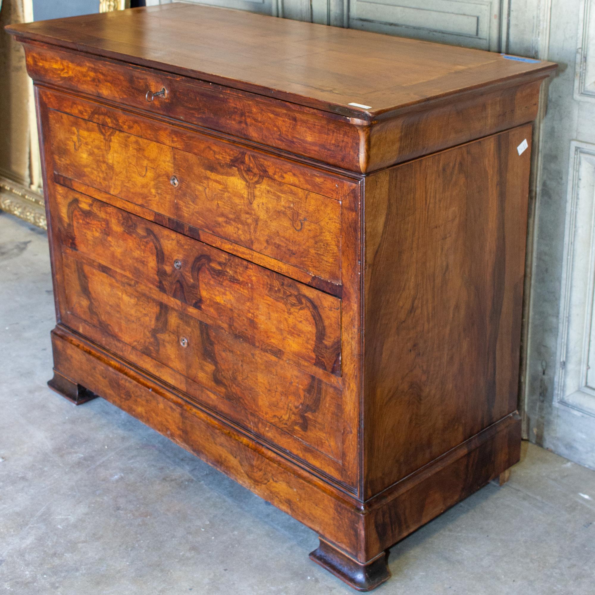This antique French Louis Philippe style chest of drawers features a beautifully crafted burled wood finish with a bold patterning. The piece has four drawers: one narrow top drawer and three deeper, lower drawers. The carved wood escutcheons are