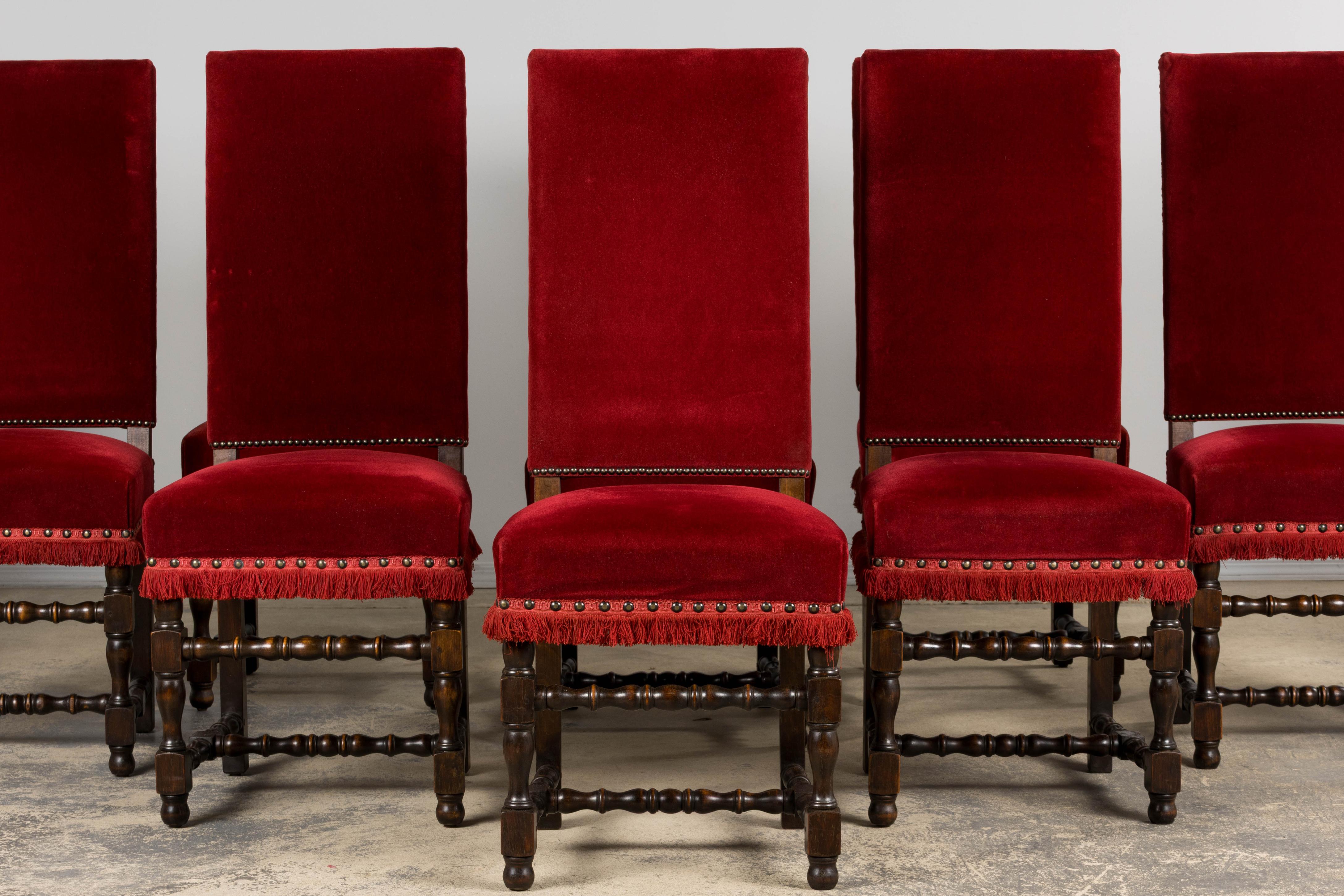 A grand set of ten early 20th century French Louis XIII style dining chairs, sourced in Provence. Each chair features a tall rectangular back and generously cushioned seat upholstered in a deep red velvet, finished with brass nailhead trim and a
