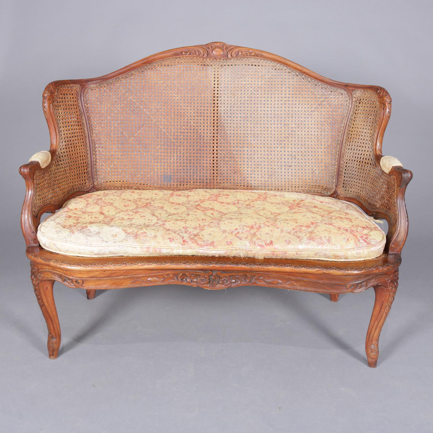 An antique French Louis XIV settee features wormwood walnut frame with carved scroll and foliate crest, scroll form arms and raised on cabriole legs, caned back, arms and seat, 19th century.

Measures: 385
