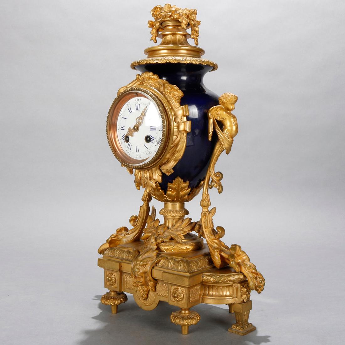 An antique French Louis XIV mantel clock offers gilt bronze and porcelain construction with cobalt blue case having foliate crest, flanking cherub handles, acanthus and foliate elements, seated on base with central lion head mask and raised on