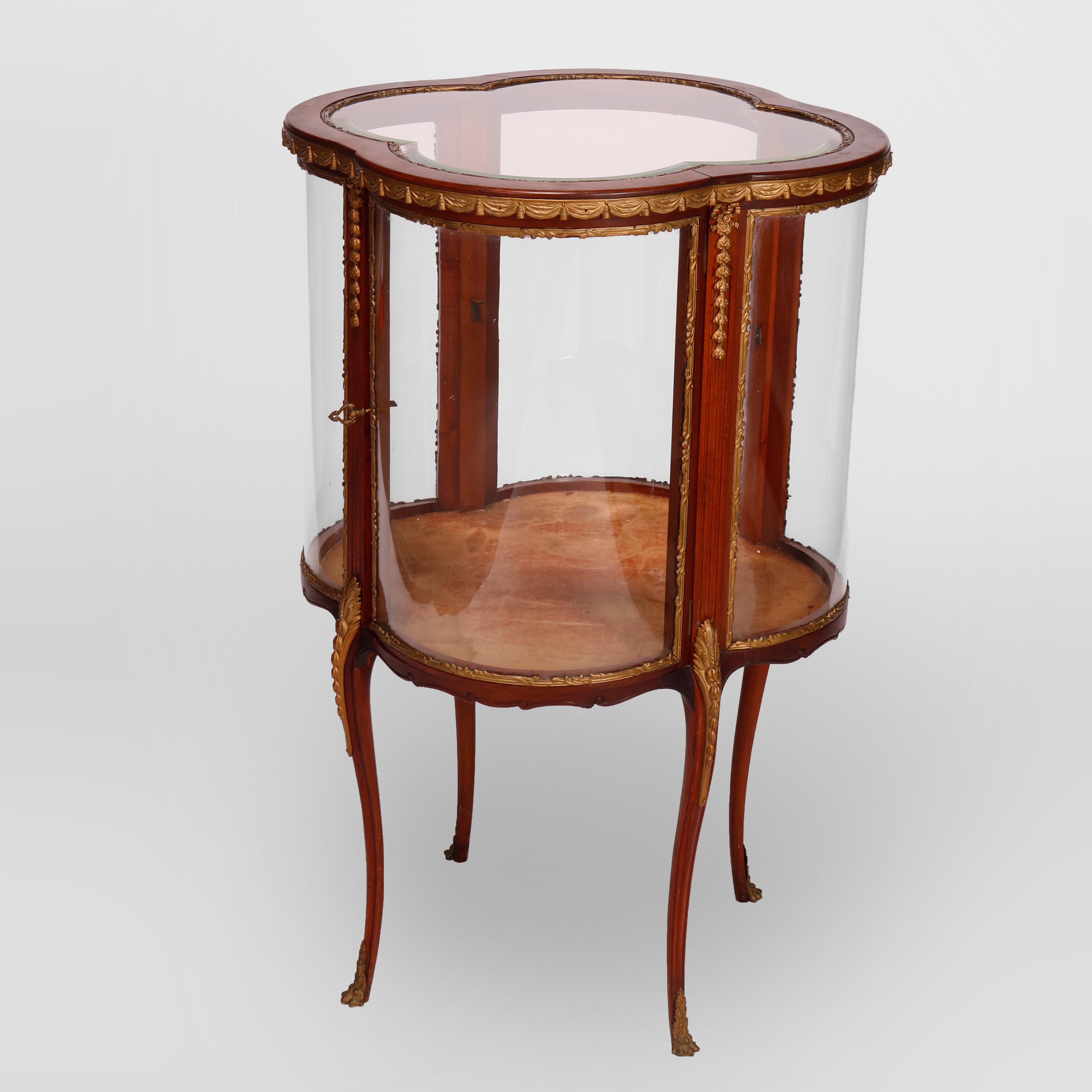 An antique French Louis XIV display vitrine offers flame mahogany frame in serpentine cloverleaf form with curved glass sides and single door, raised on cabriole legs, cast ormolu in foliate, scroll, inverted bellflower, drape and tassel form